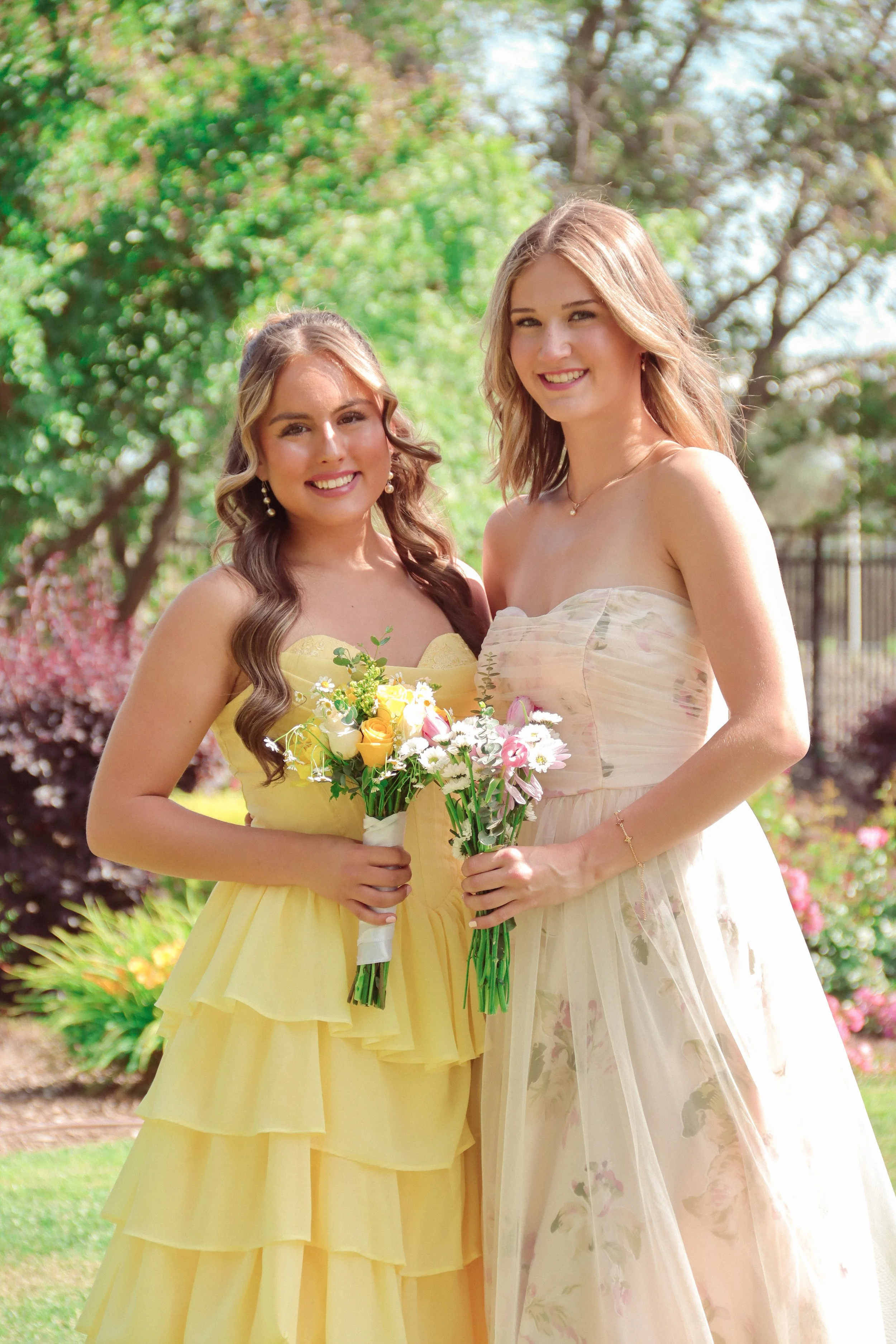 Two women in pastel-colored dresses standing outdoors with trees and greenery in the background, holding bouquets of flowers and smiling.