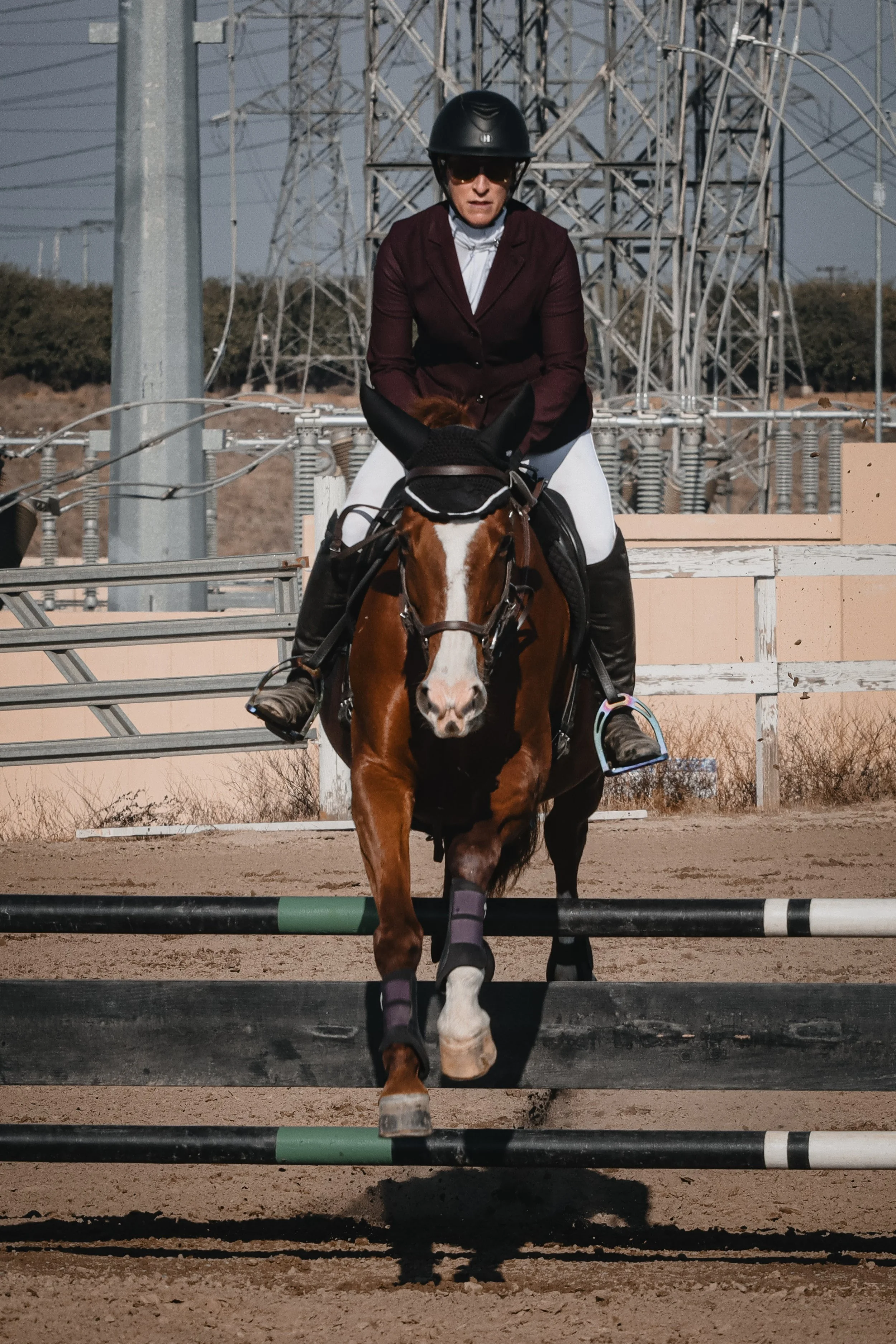 A woman riding a brown and white horse, wearing a helmet and riding attire, crossing a jumping obstacle in an outdoor equestrian arena with electrical towers in the background.