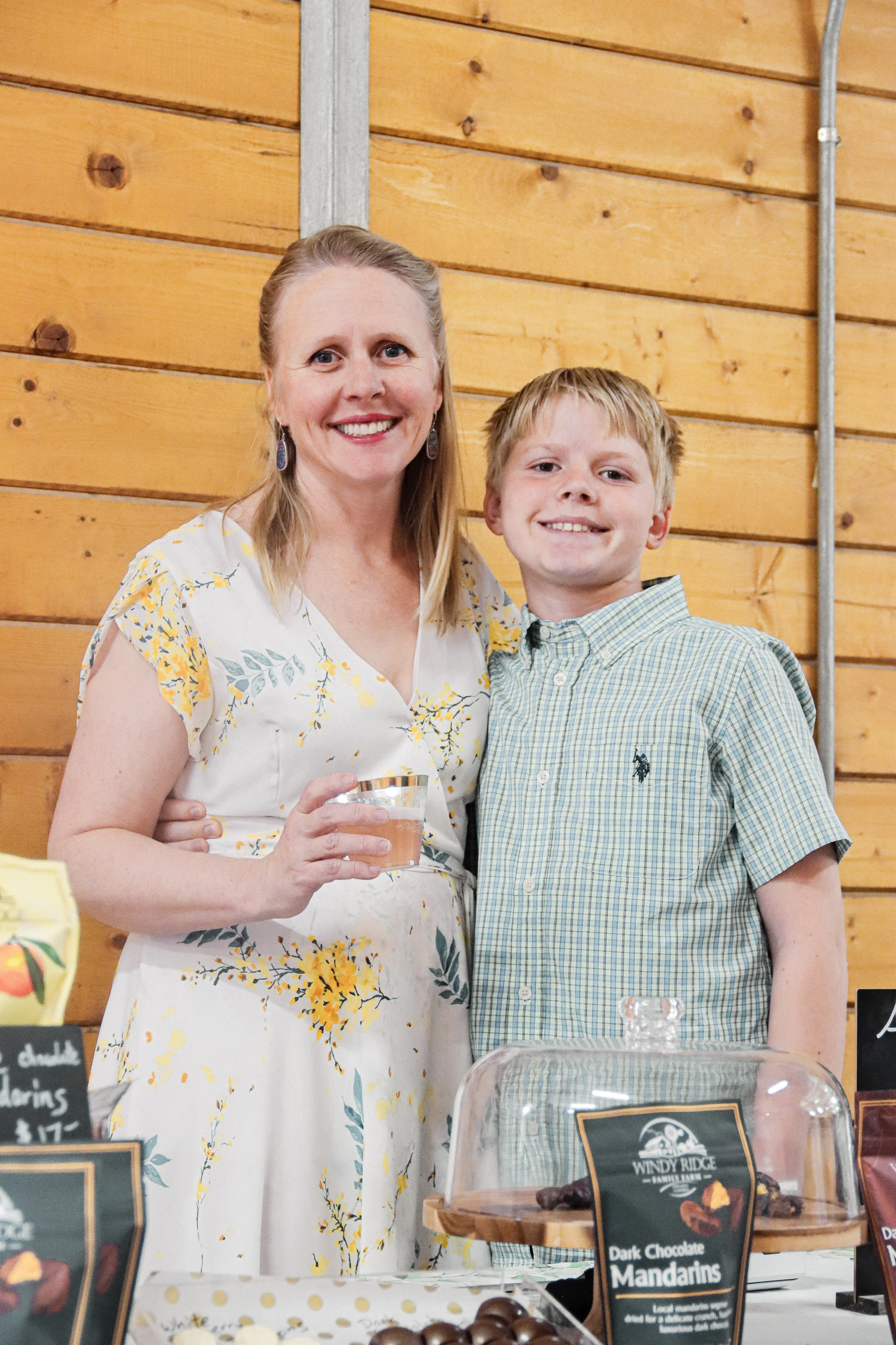 A woman and a young boy standing together at a market stall with chocolate confections and signs for dark chocolate mandarins.
