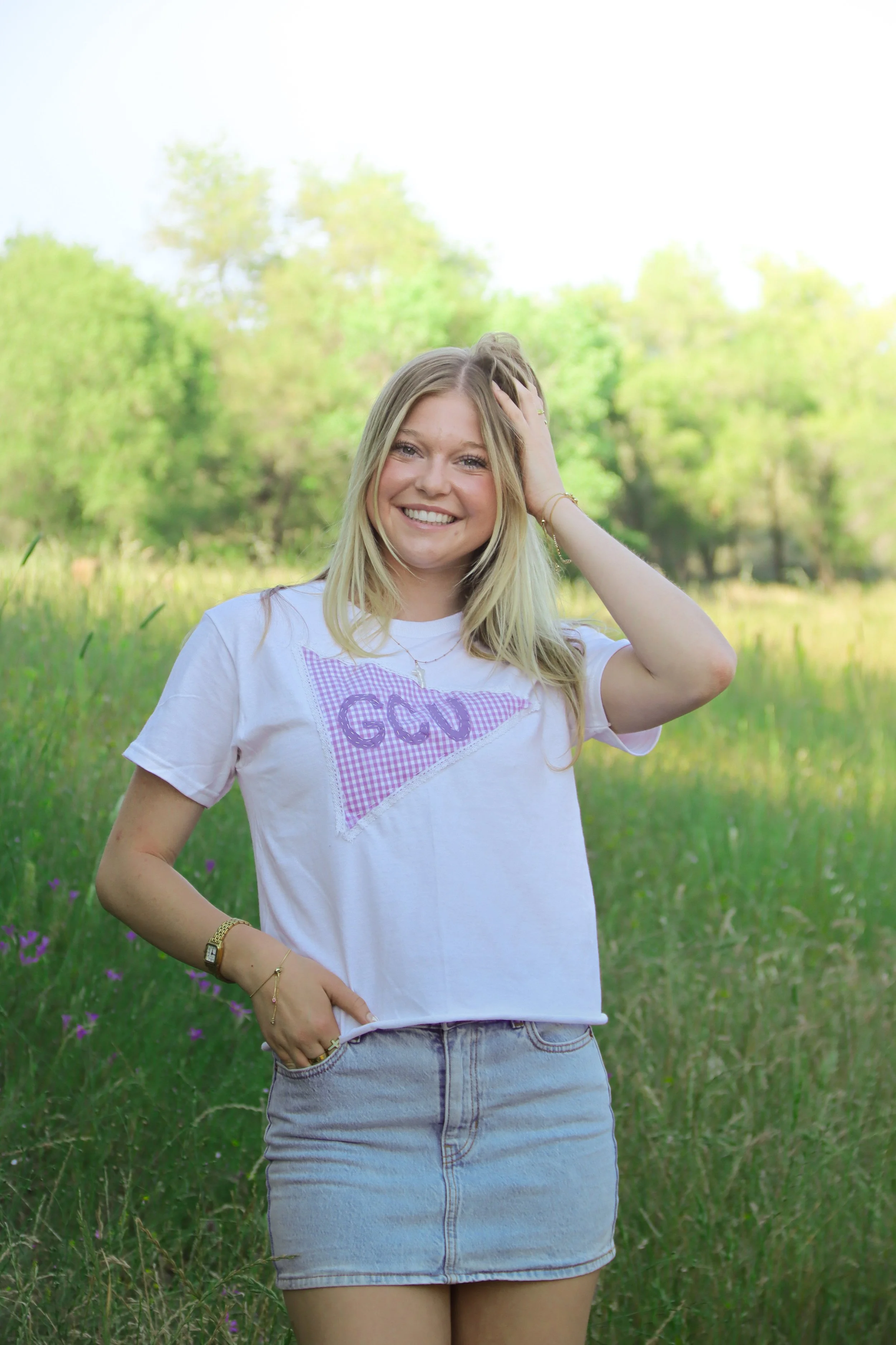A young woman with blonde hair smiling outdoors in a green, grassy field with trees in the background, wearing a white t-shirt and denim skirt.