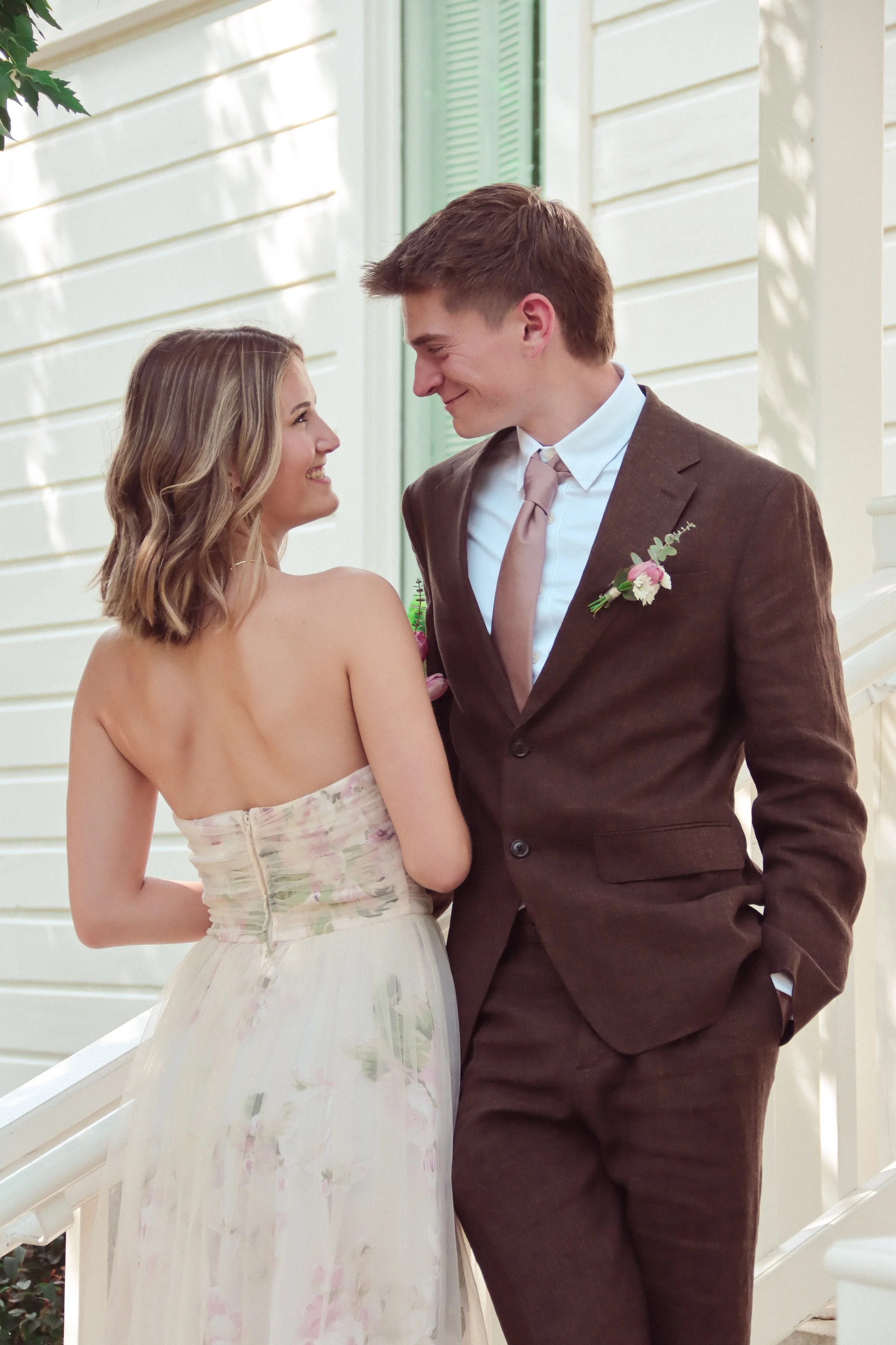 A young couple with light skin and average physiques standing close together on a staircase, smiling and gazing at each other. The woman has shoulder-length blonde hair and is wearing a strapless dress with a floral pattern and a zipper at the back. 