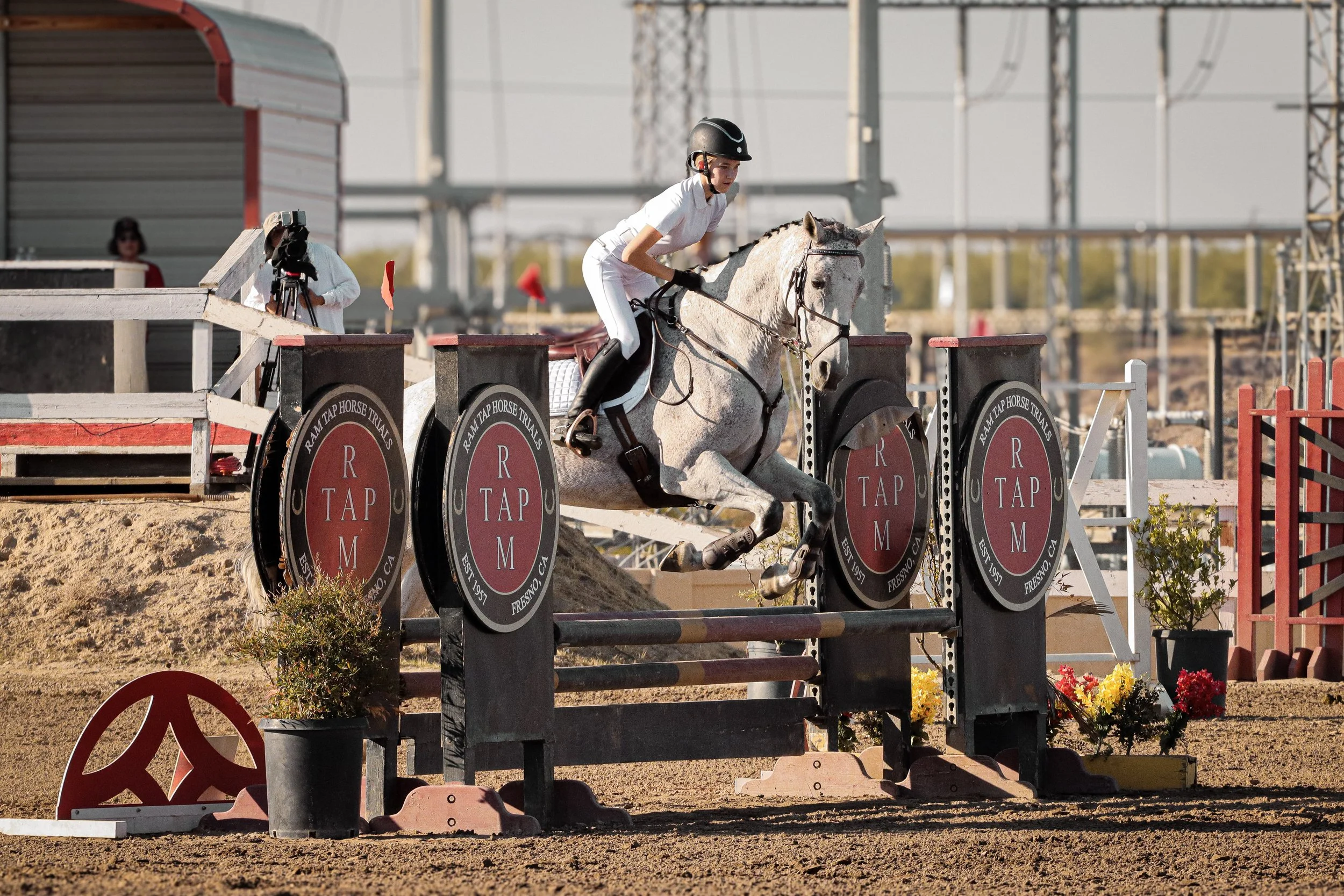 A female equestrian wearing a helmet and white riding outfit riding a gray horse over a show jumping obstacle outdoors, with a photographer capturing the moment nearby and various banners and equipment around.