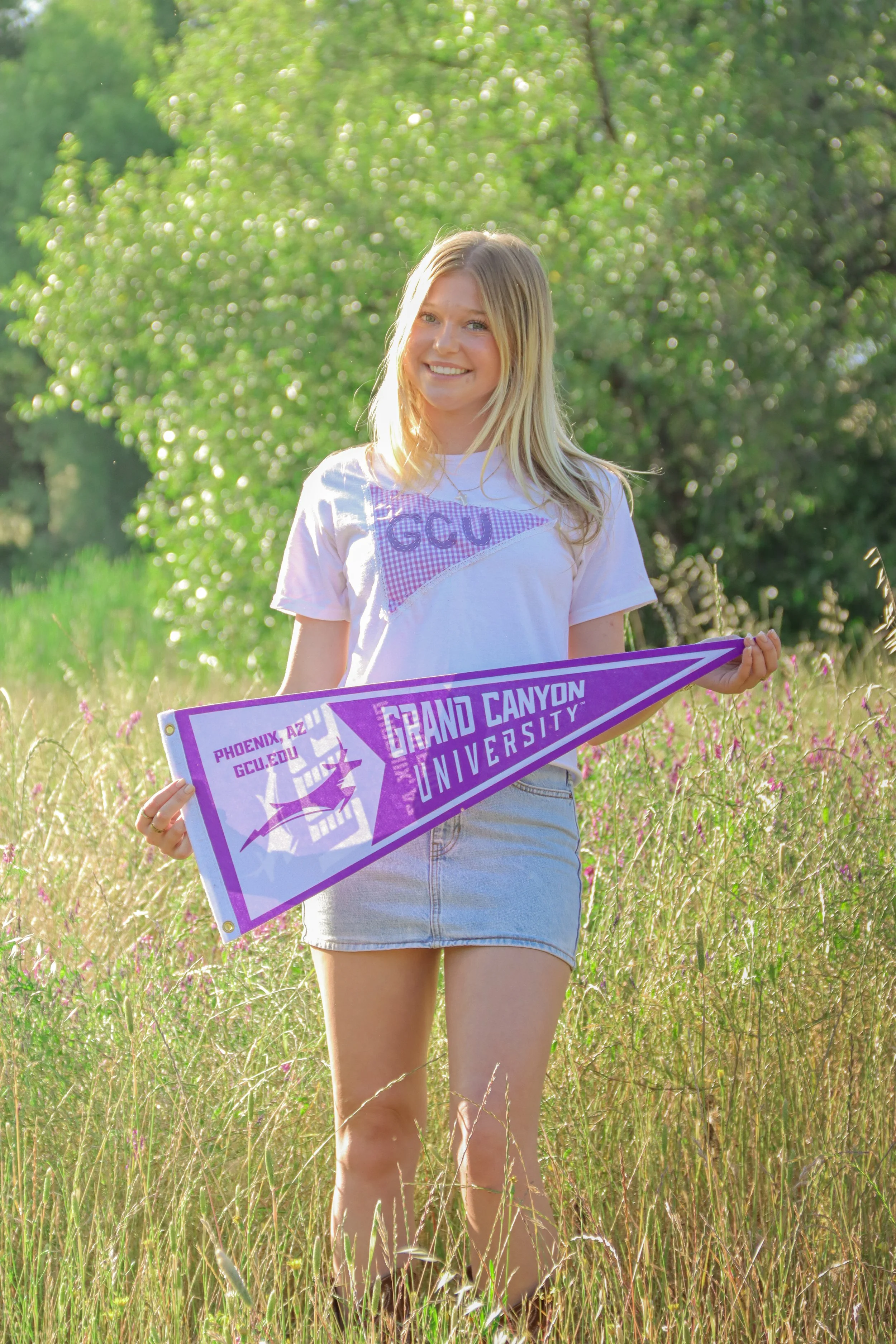 A young woman with blonde hair smiling outdoors in a field of tall grass and wildflowers, holding a purple and white pennant that reads "Grand Canyon University." She is wearing a white T-shirt and light-colored denim skirt.