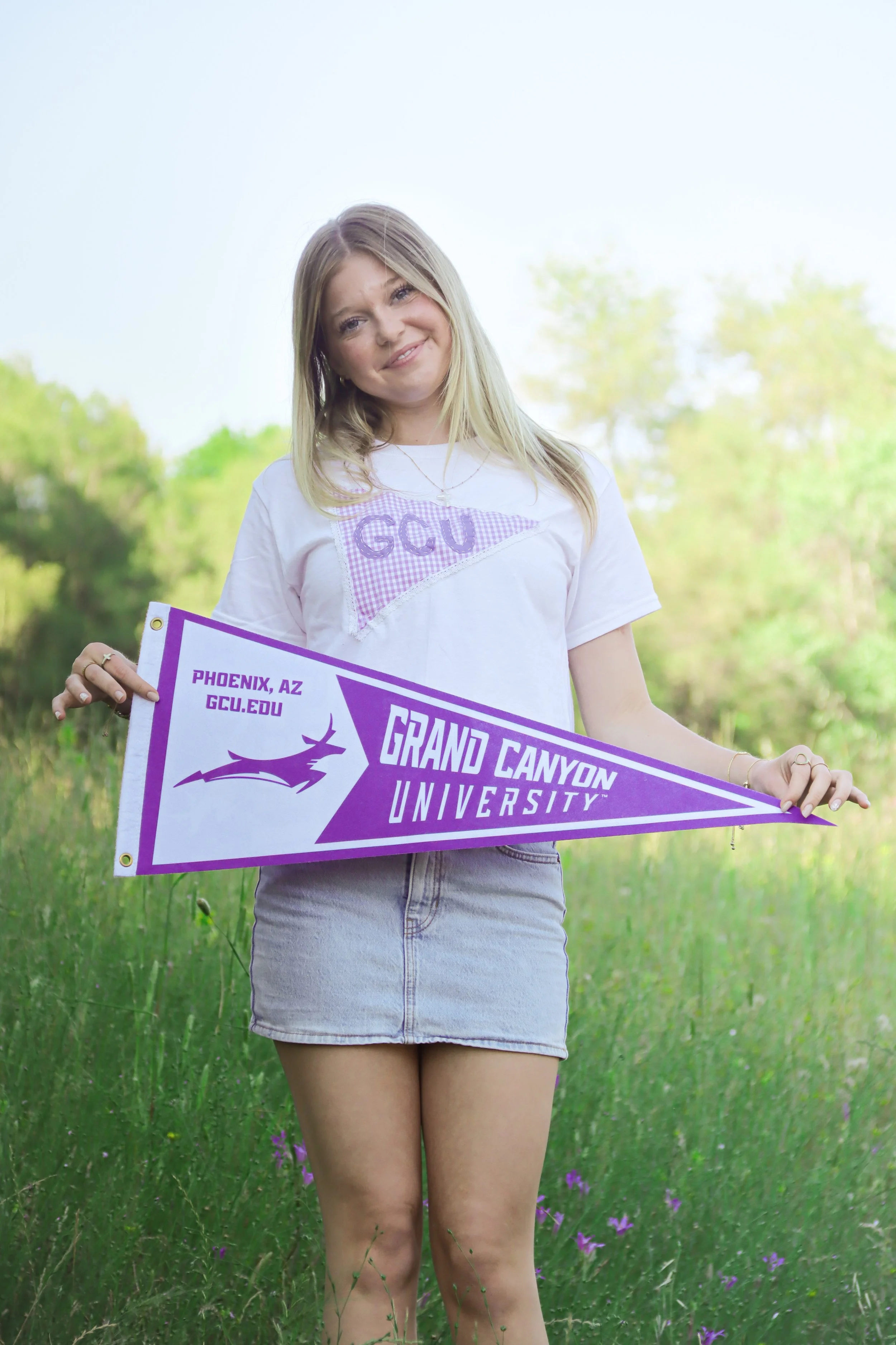A young woman outdoors holding a purple and white Grand Canyon University pennant, smiling, with trees and grass in the background.