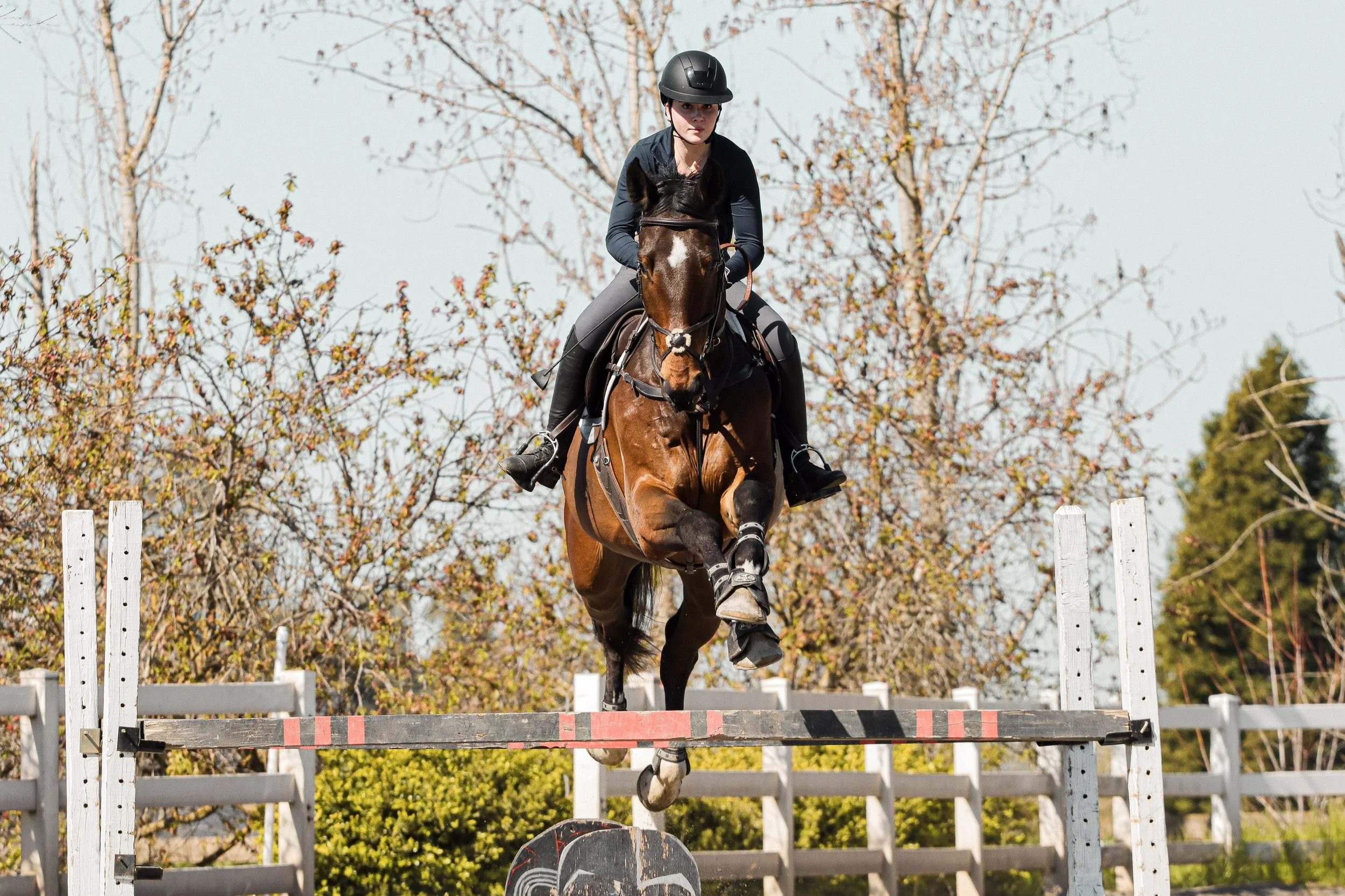 A young woman wearing a helmet riding a horse over a jump during an outdoor equestrian event.