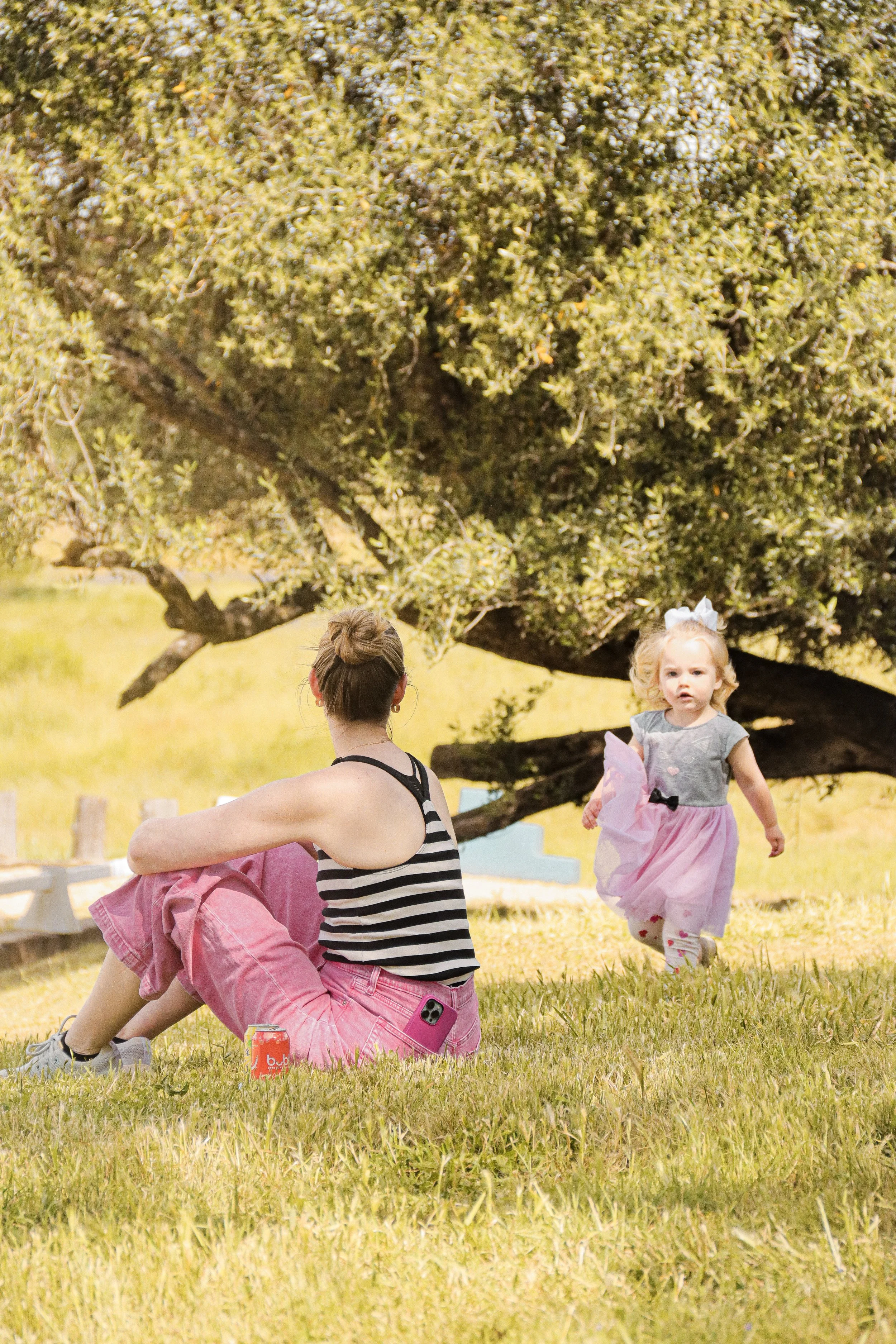 A woman sitting on the grass with a young girl running towards her outdoors, with trees and a fence in the background.