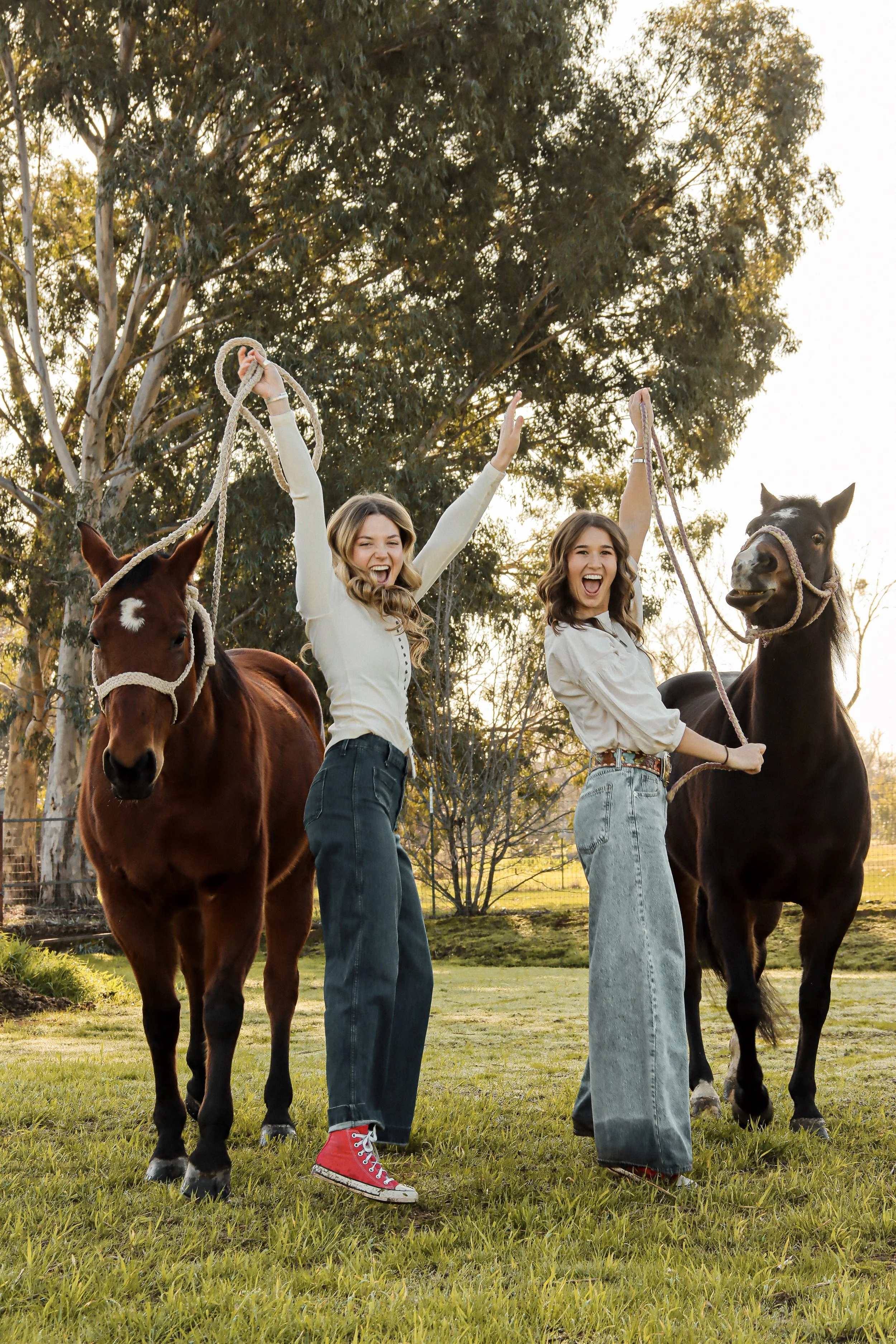 Two smiling young women in casual clothing with their arms raised, standing between two horses in a grassy outdoor setting with trees in the background.