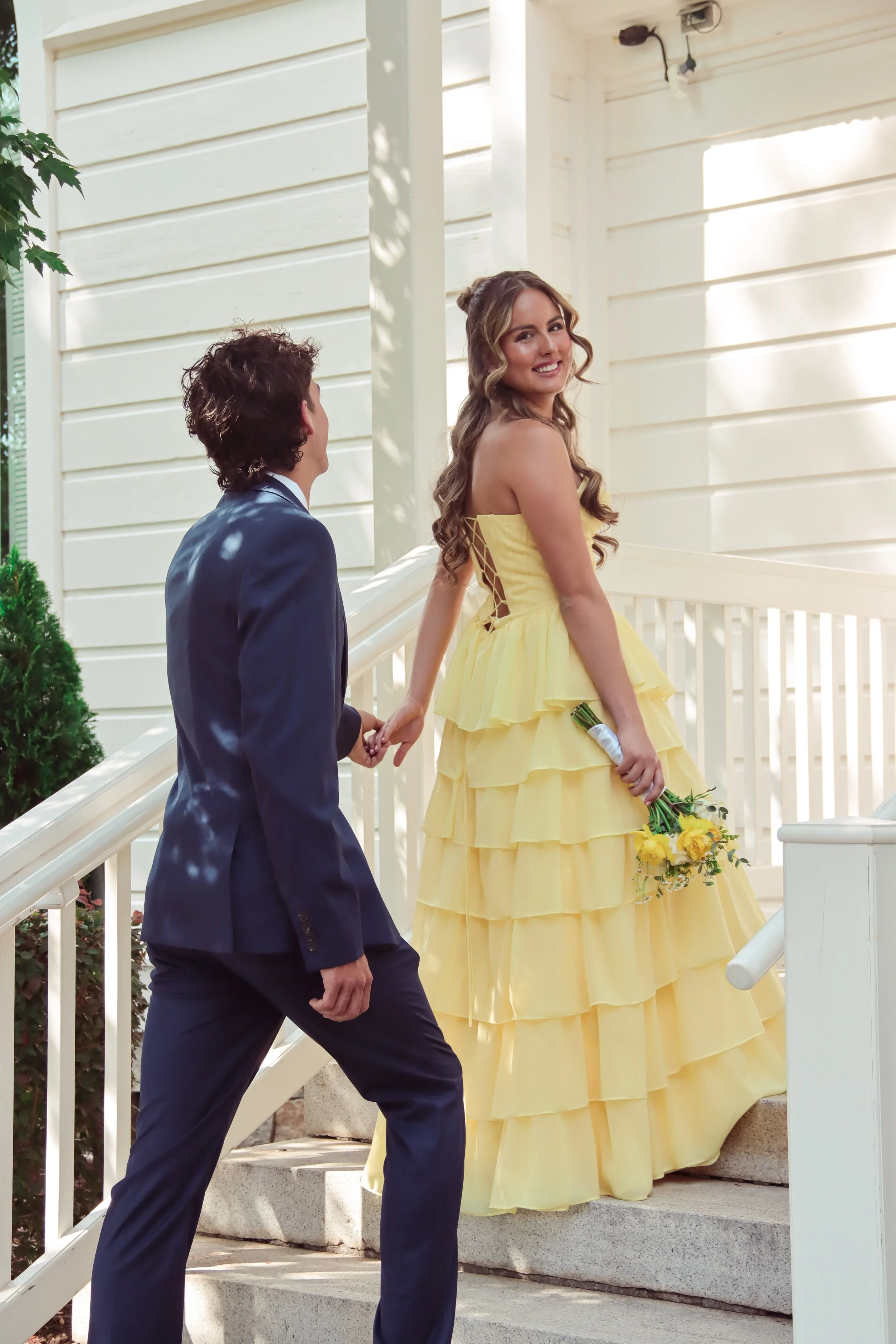 A woman in a yellow dress holding a bouquet of yellow flowers is standing on a porch, looking happily over her shoulder. A man in a dark suit is holding her hand and looking at her. The porch has white wooden railings and steps.