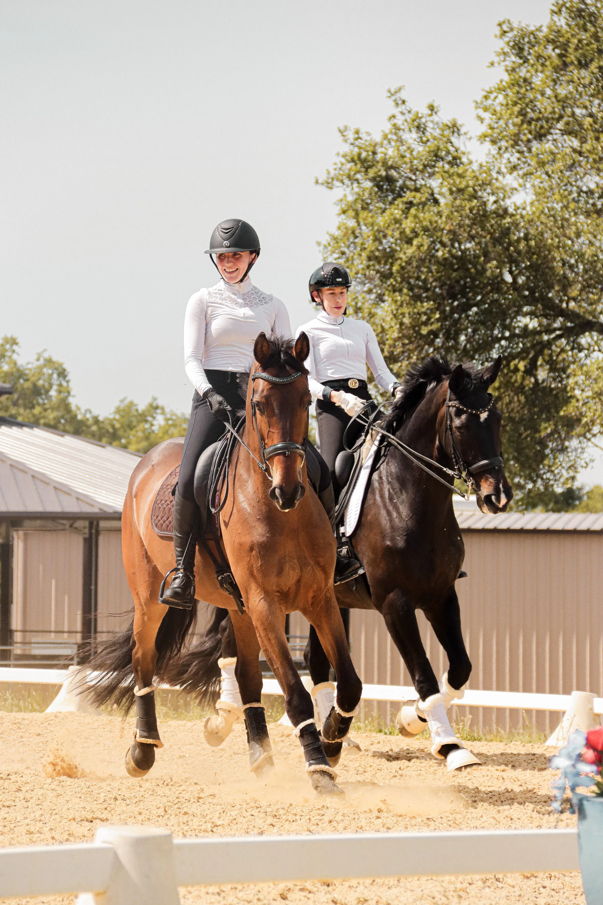 Two women riding horses in a dressage arena, both wearing helmets and white shirts, with trees in the background.