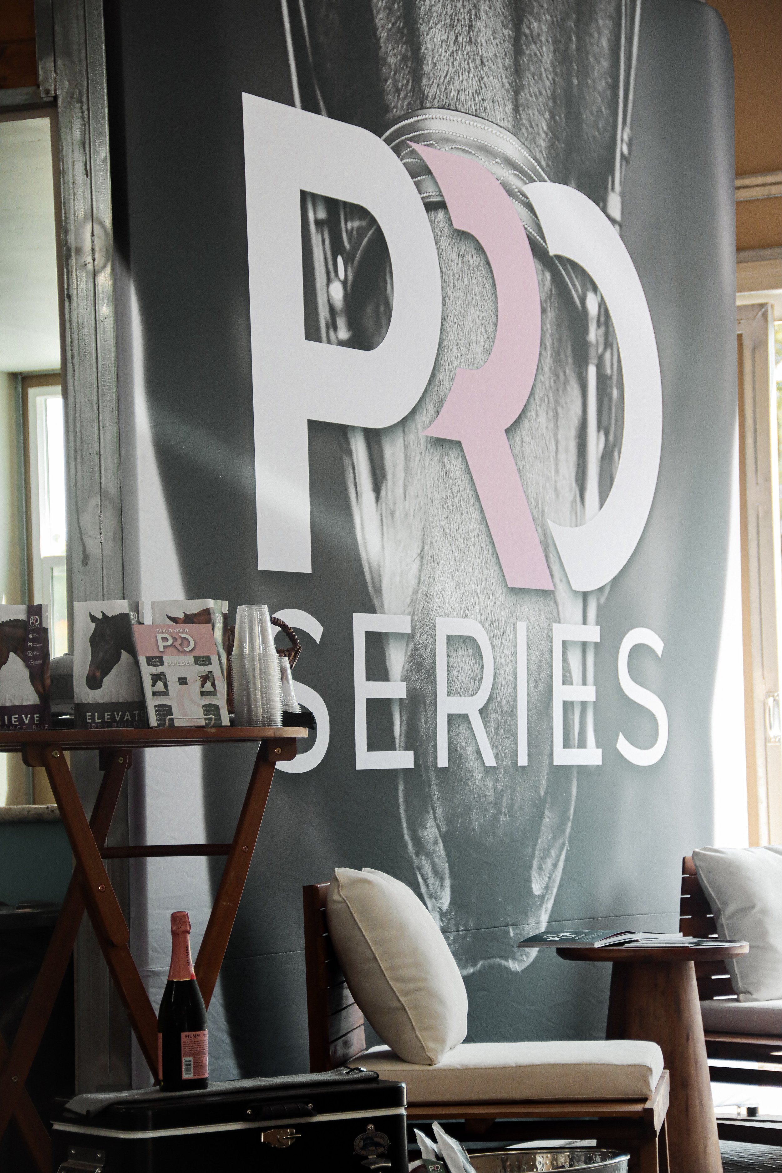Indoor scene with a large banner display showing the letters 'PD' and the word 'SERIES'. A small table with miscellaneous items including a bottle with a pink label, a stack of clear plastic cups, a magazine or booklet, and a cushioned chair with a b