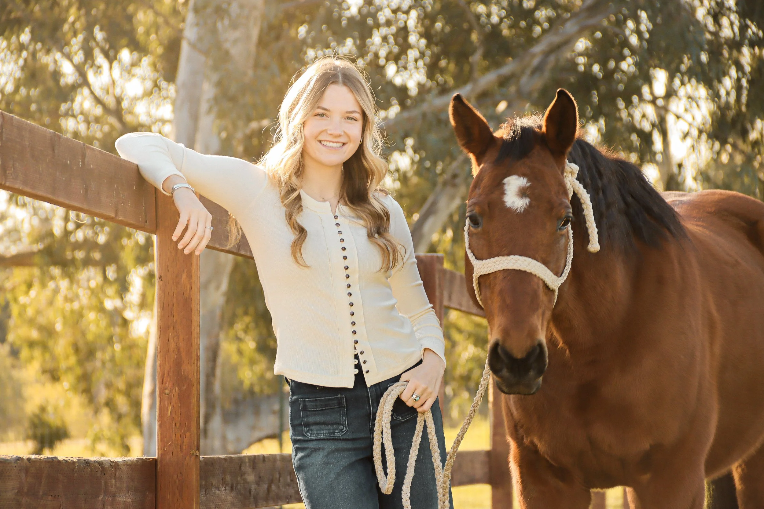 A young woman with wavy blonde hair smiling and leaning on a wooden fence next to a brown horse with a white star-shaped marking on its forehead, outdoors with trees in the background during golden hour.
