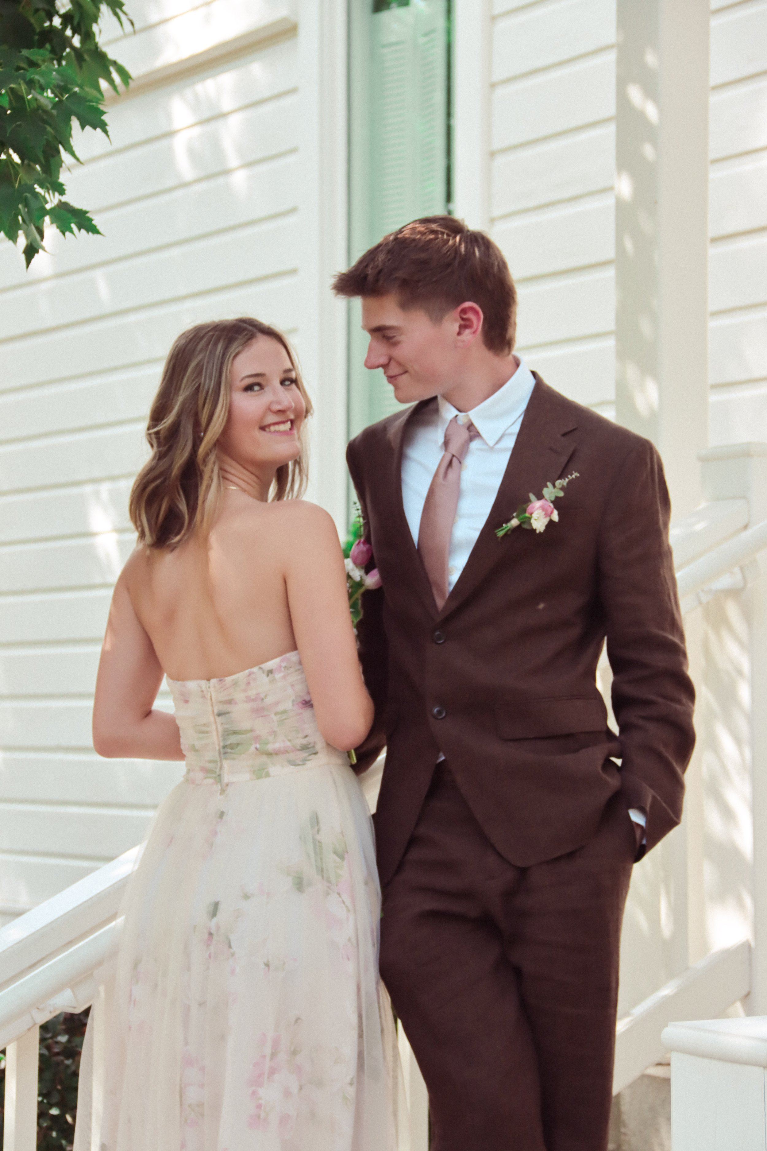 A happy couple dressed for a wedding, standing close together next to a white railing outside, with a white house and green foliage in the background.