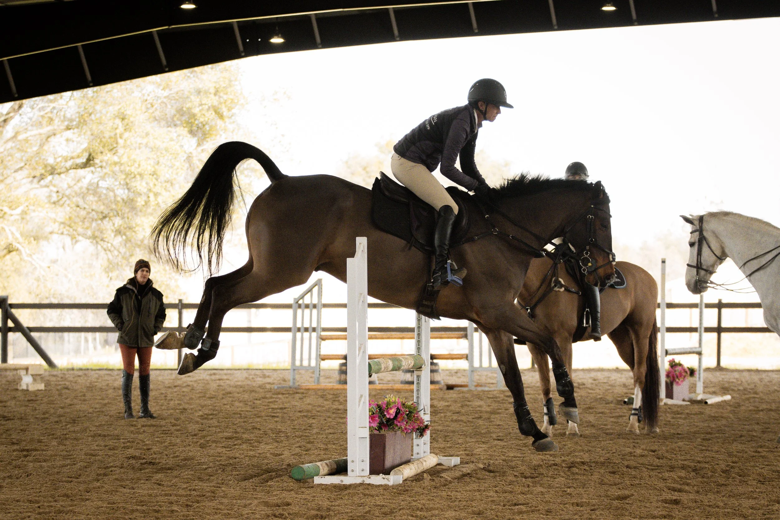 A person riding a horse over a jump in an indoor equestrian arena, with another person watching nearby.