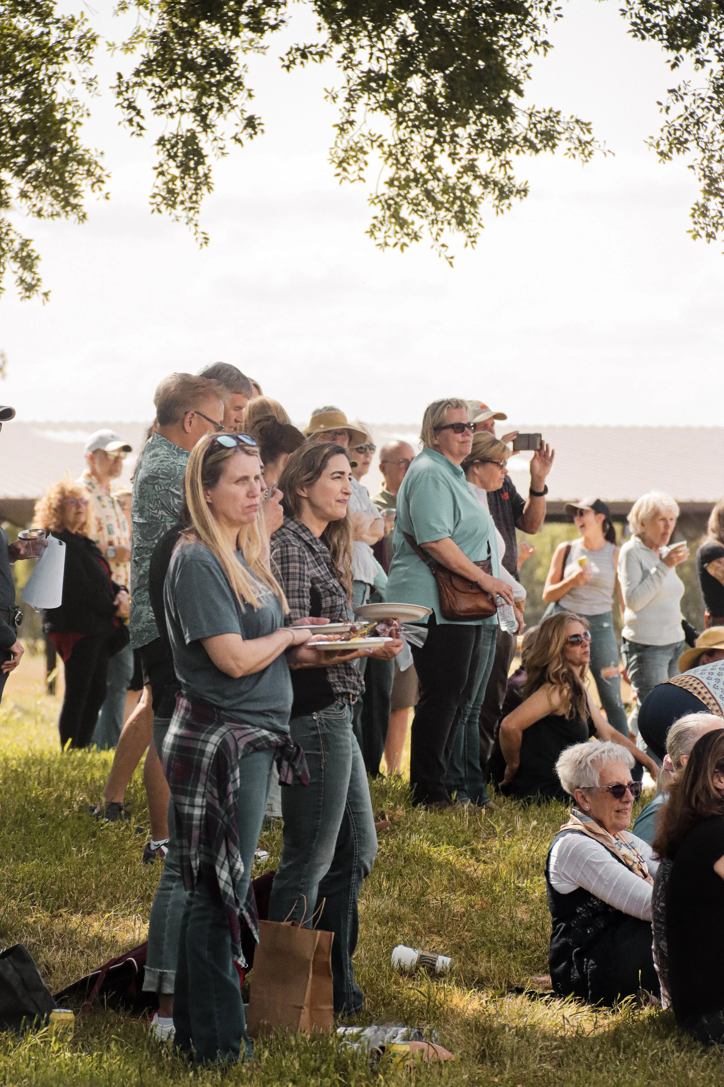 A crowd of people gathered outdoors, some standing and some sitting on the grass, during daytime with trees overhead.