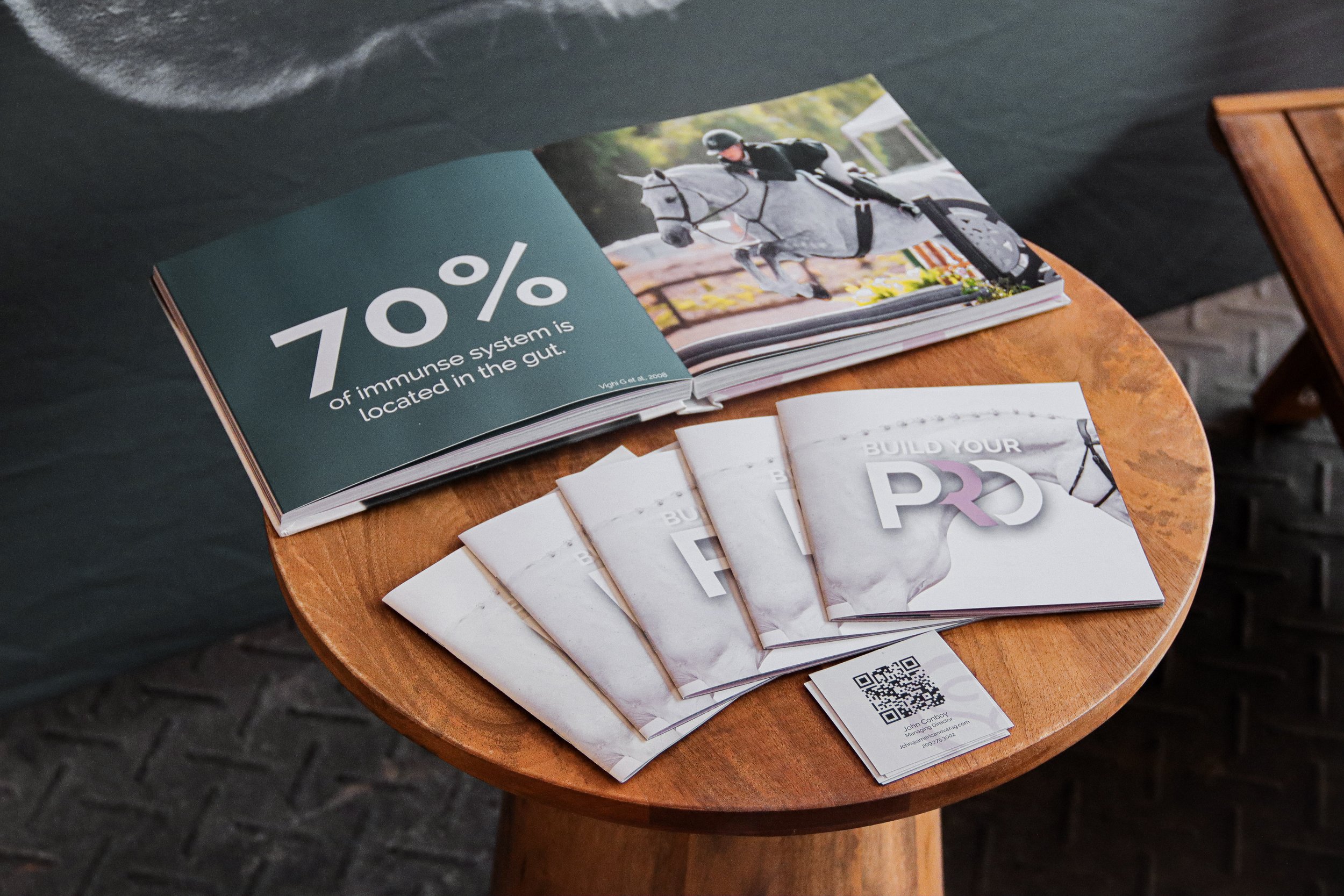 Open magazine on a wooden table showing a picture of a person riding a white horse and a statistic stating 70% of immune system is located in the gut. Brochures and business cards are also on the table.