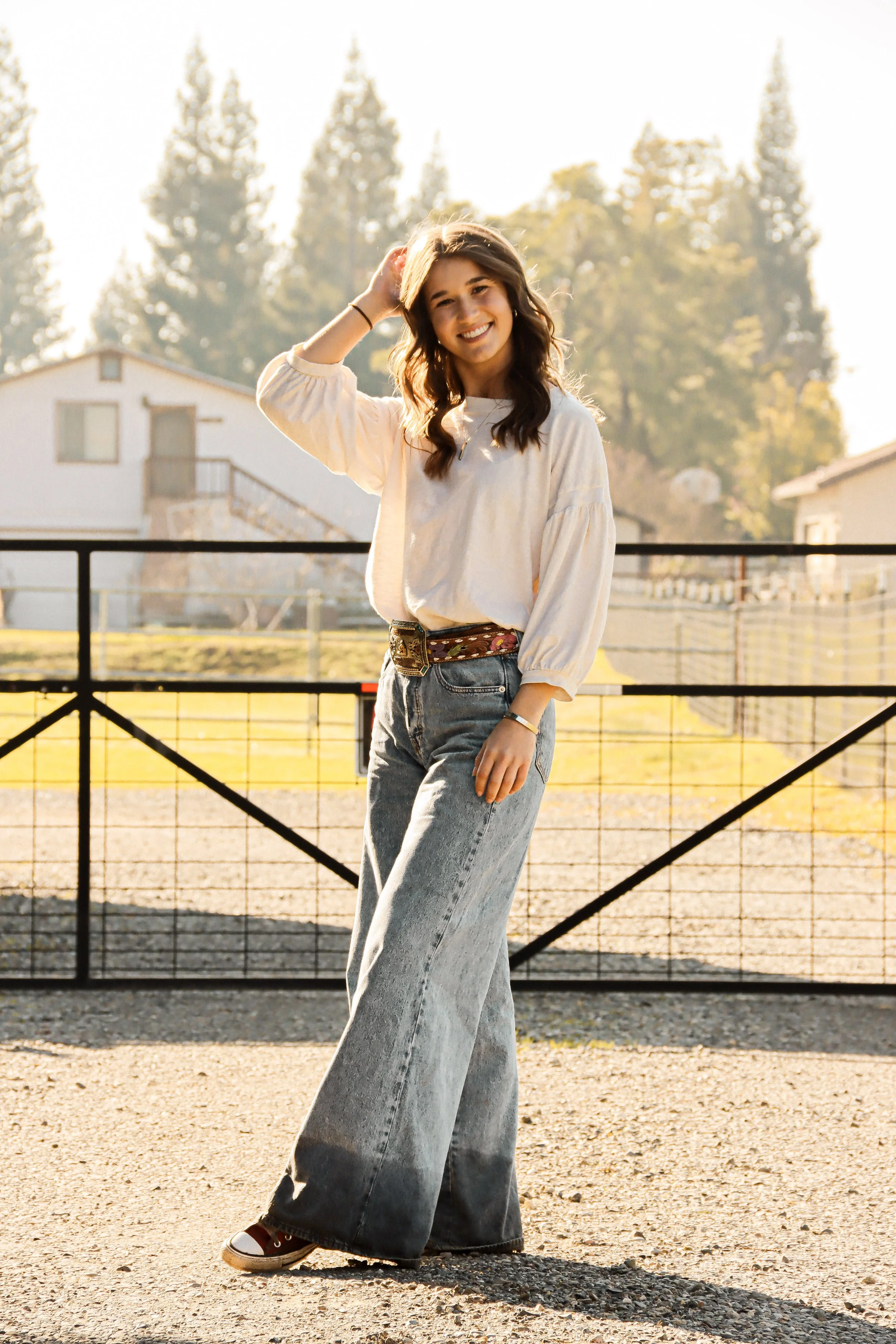 Young woman with long wavy hair smiling, wearing a light-colored blouse, high-waisted wide-leg jeans, and sneakers, standing by a black gate outdoors in the evening sunlight.