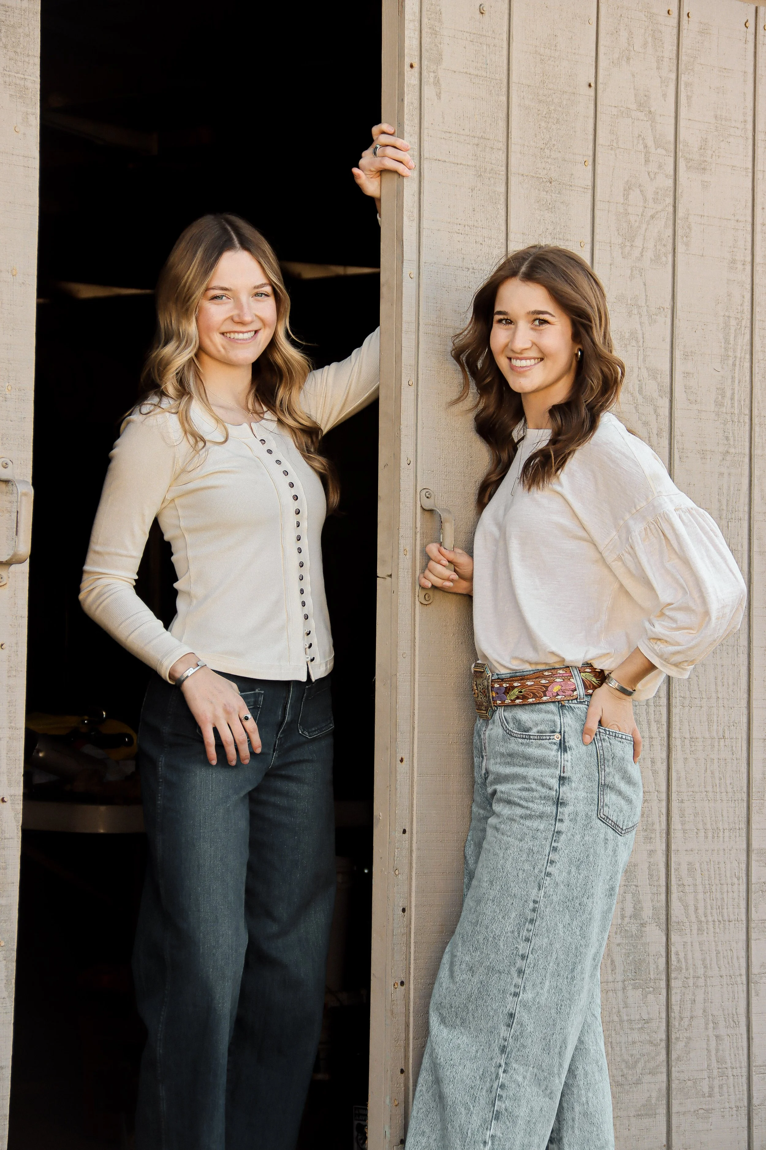 Two young women smiling and standing by the open door of a barn or shed on a farm, dressed casually in jeans and long-sleeve shirts.