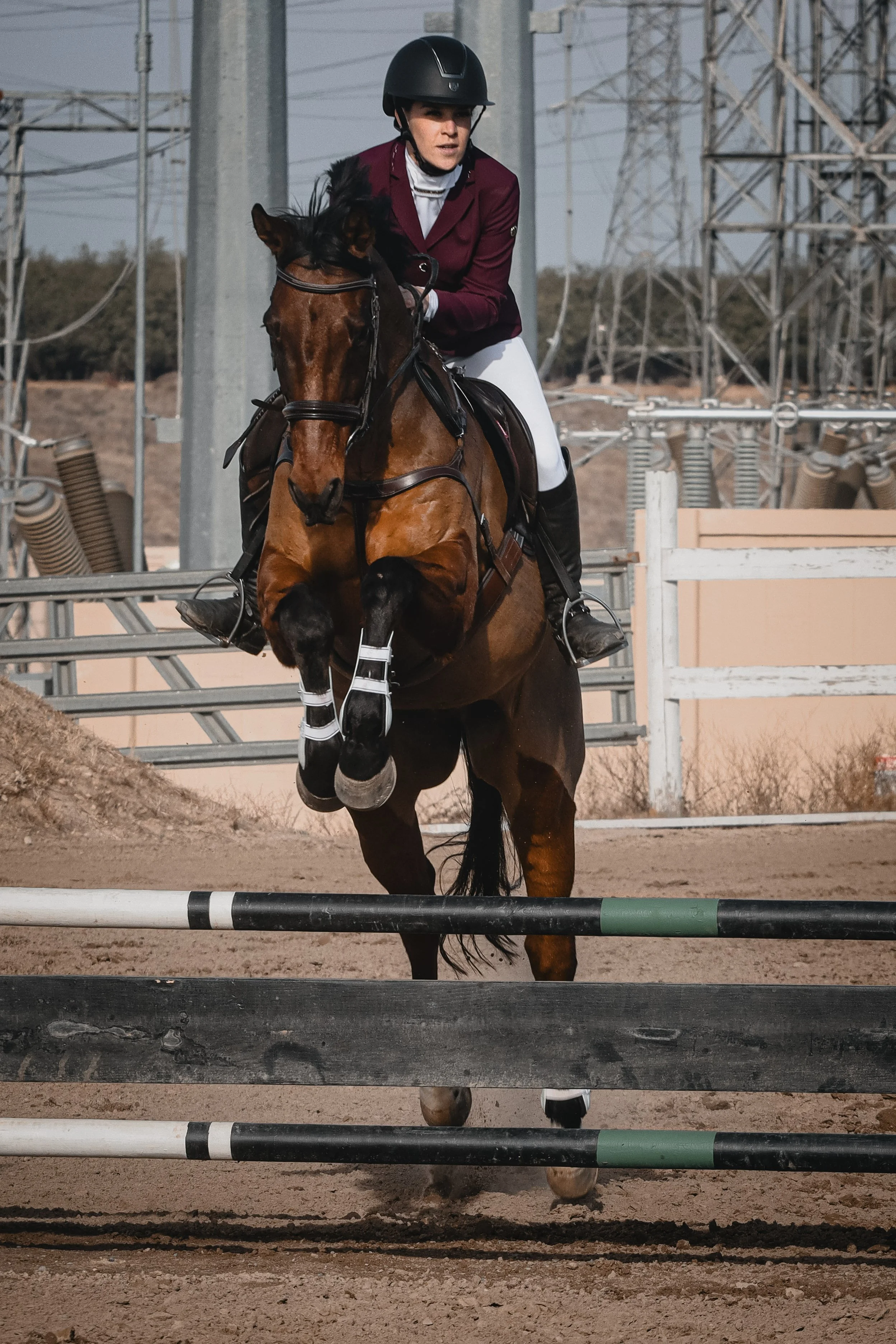 A female equestrian rider in a maroon jacket, white breeches, and black riding helmet guides a brown horse over a show jumping obstacle on a dirt arena with electrical towers and wires in the background.