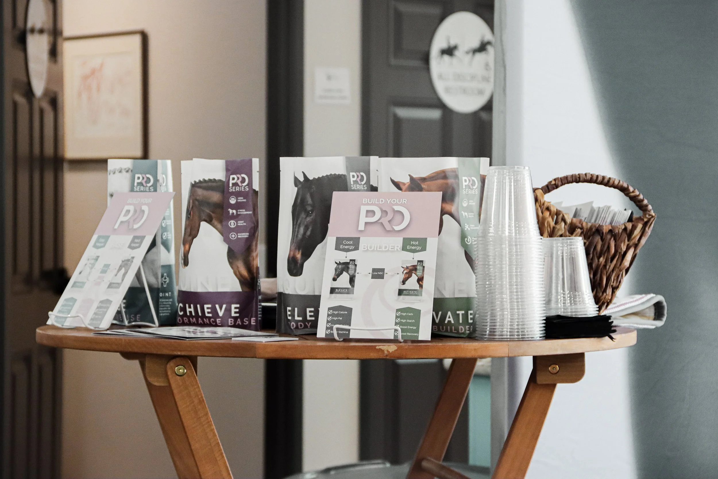 A display of horse-themed nutritional supplements and promotional materials on a small round wooden table with stacks of clear plastic cups. The background shows a door with a circular sign featuring horses and a faintly visible painting on the wall.