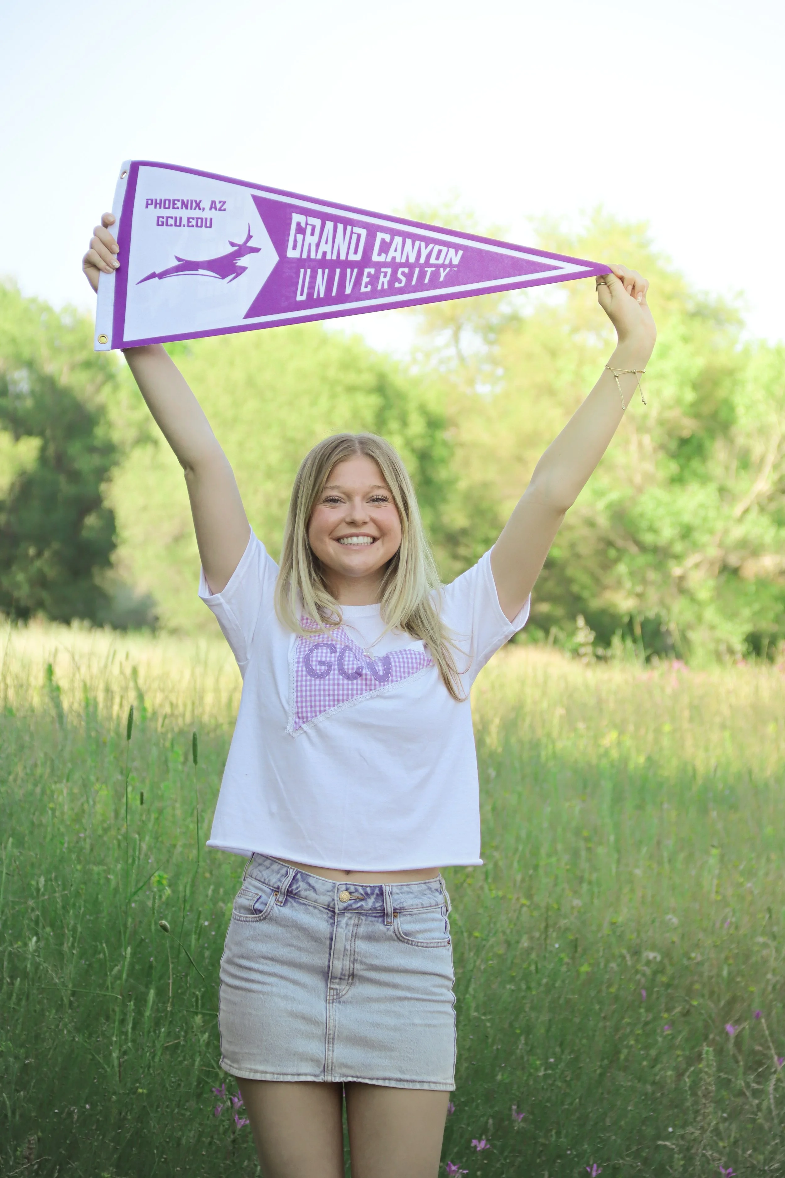 A girl standing in a grassy field holding a purple and white pennant that reads "Grand Canyon University" with a logo and location "Phoenix, AZ". She is smiling, wearing a white T-shirt and a denim skirt, with a bracelet on her right wrist and a neck
