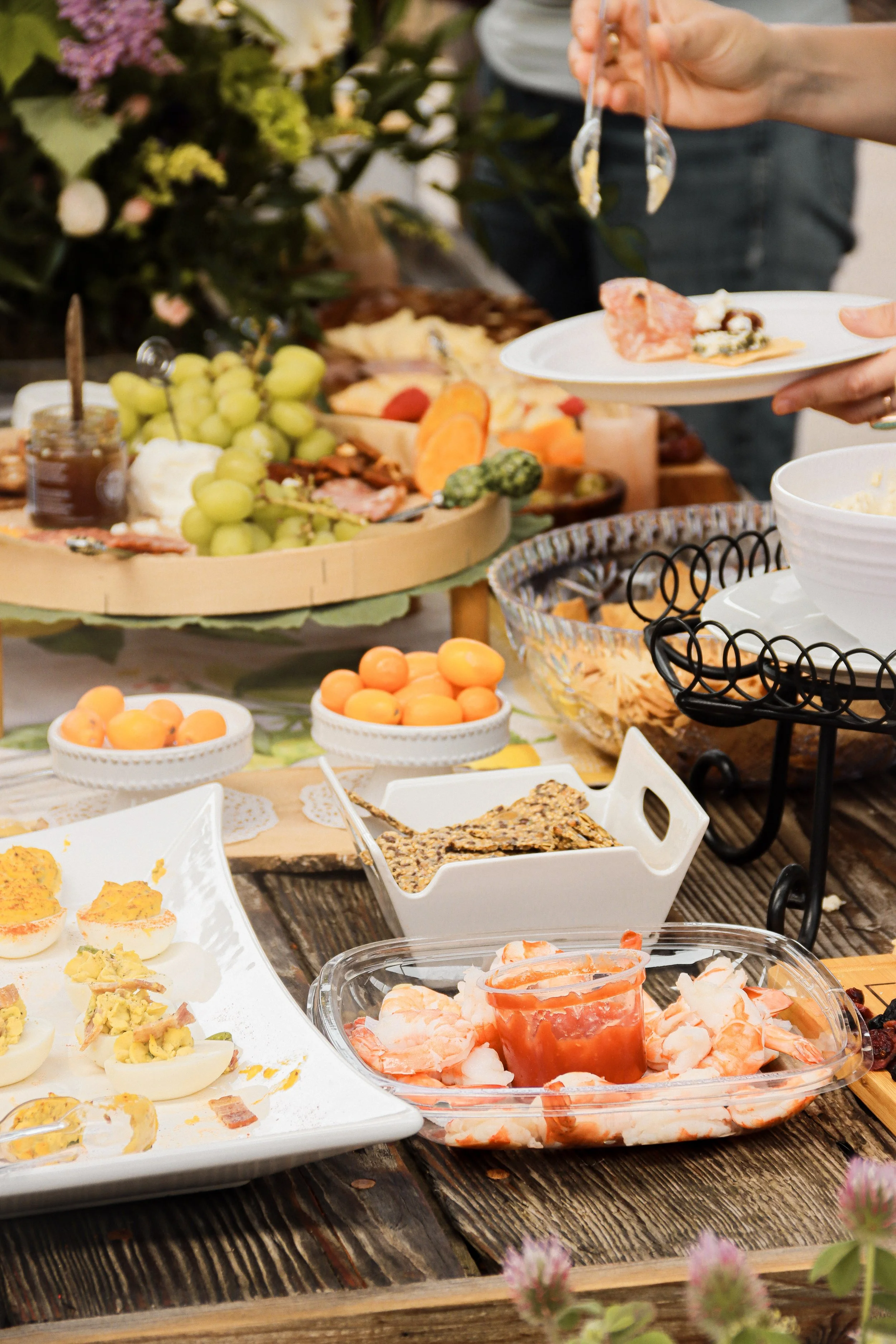 A buffet table with various dishes, including Deviled eggs, shrimp cocktail, grapes, and other appetizers, with a person serving themselves.