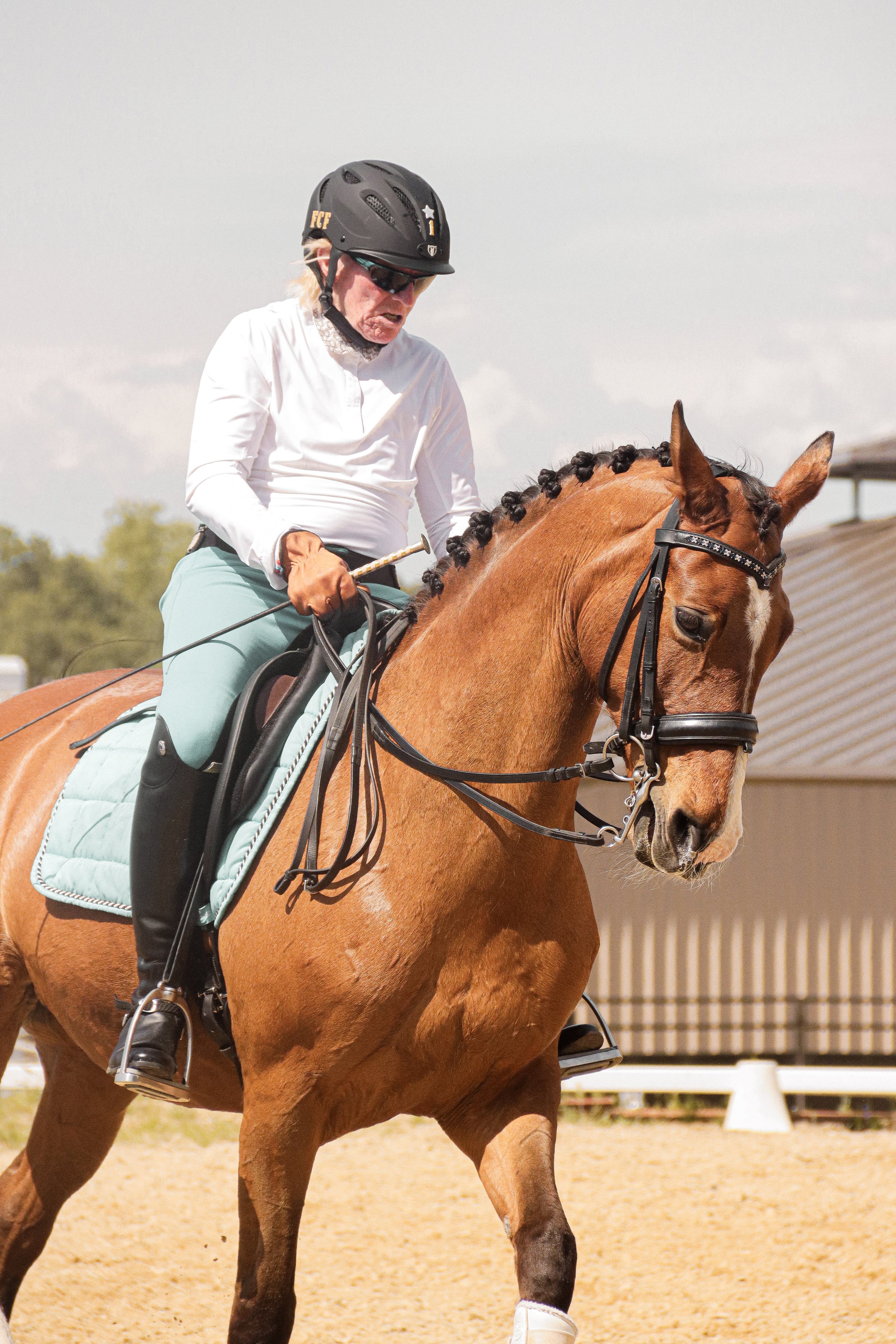 A person riding a brown horse during a daytime equestrian event, wearing a helmet, sunglasses, a white shirt, and light blue riding pants.