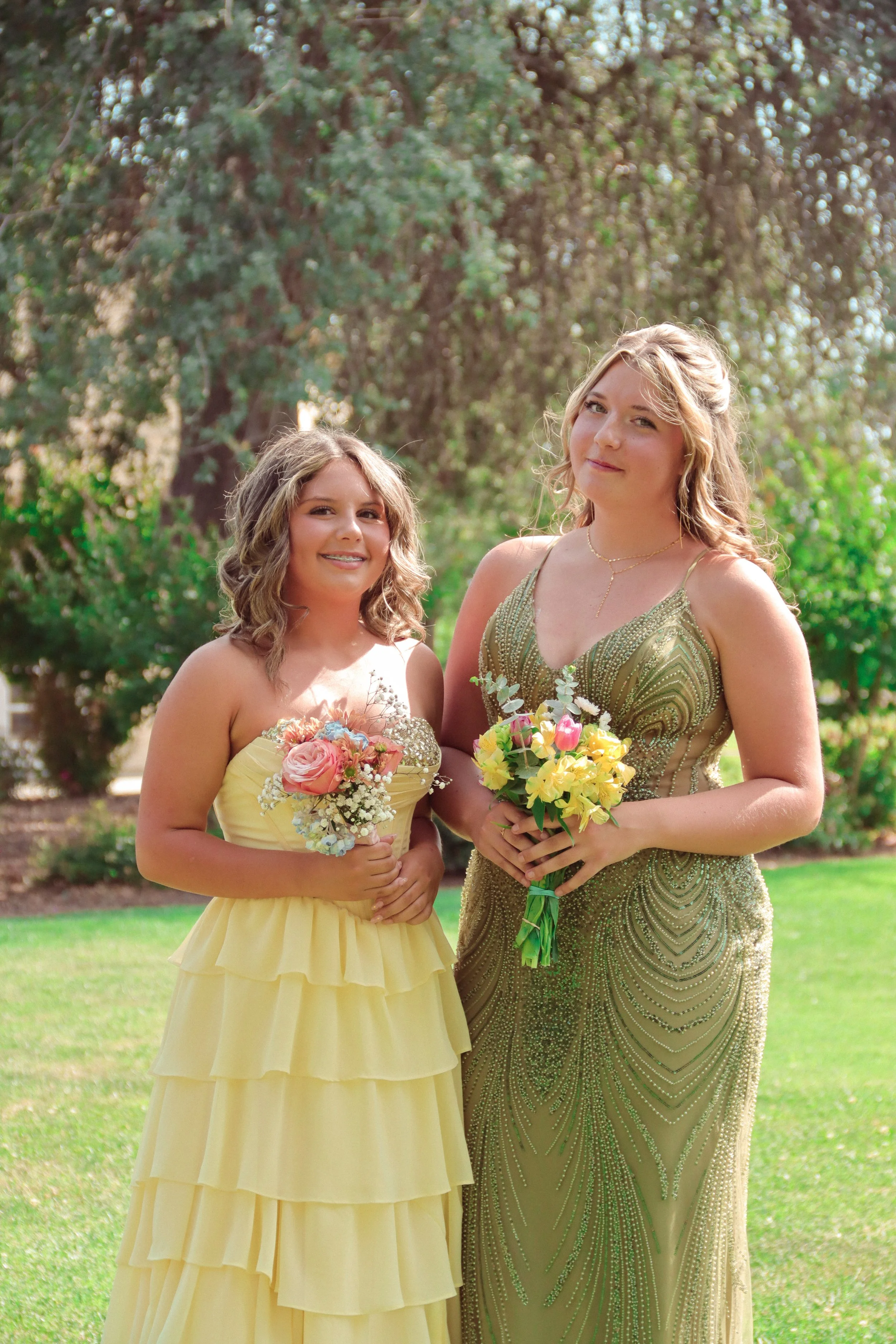 Two women in elegant dresses holding bouquets outdoors with trees and grass in the background.