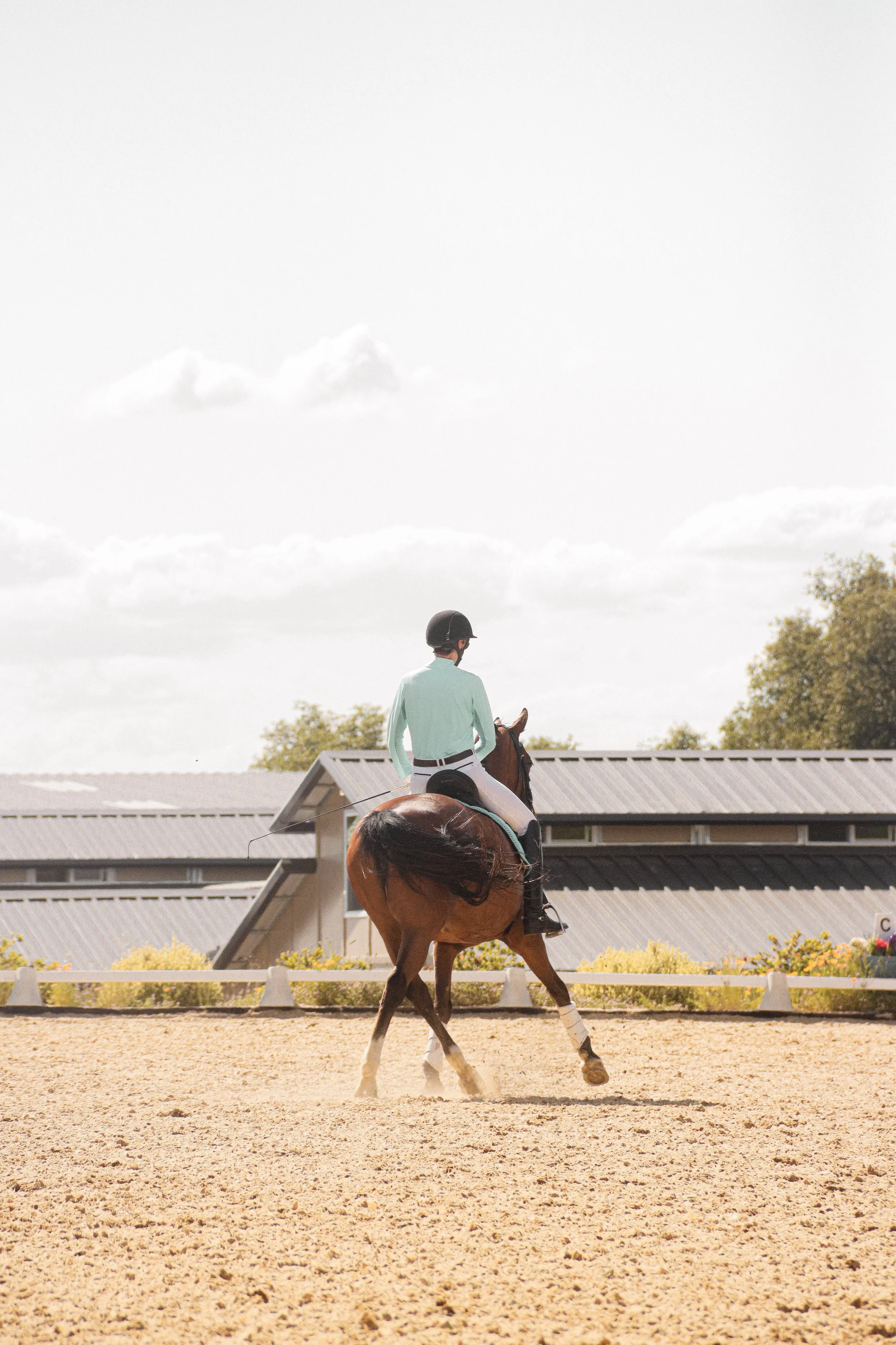 A person riding a brown horse in an outdoor riding arena during daytime, with another rider in the background wearing a helmet and a light green jacket, white riding pants, and black boots, with trees and a building with a sloped roof in the backgrou