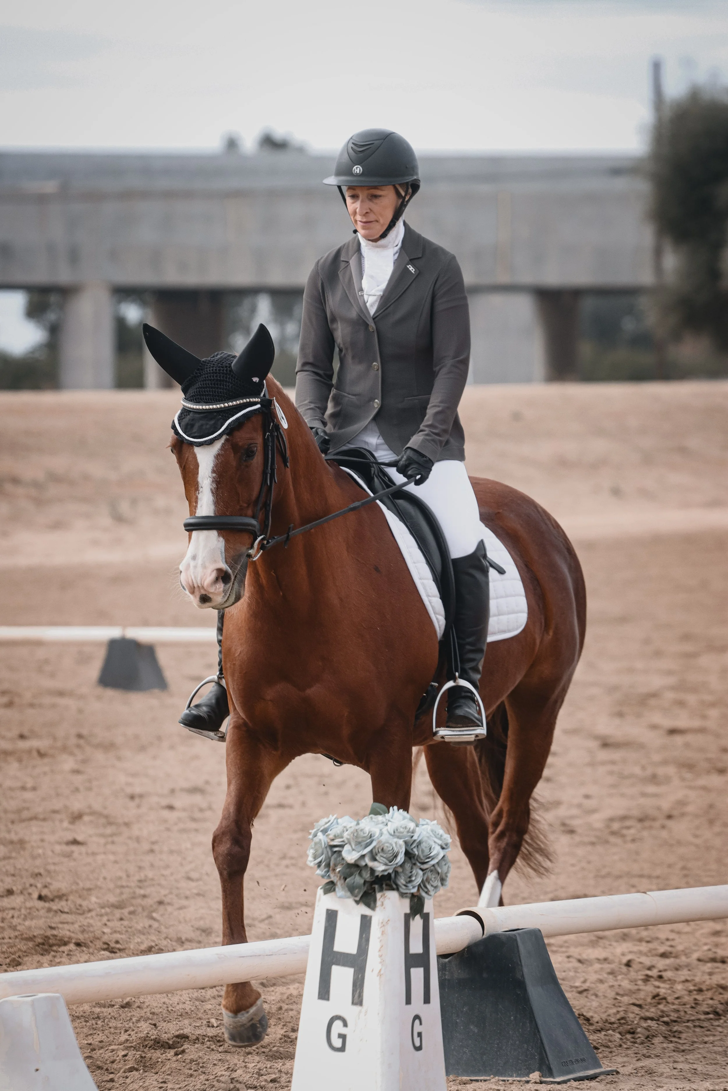 A woman in riding attire and helmet riding a chestnut horse in an outdoor arena. The horse is decorated with a black ear bonnet and a floral arrangement on its bridle. The woman is walking the horse past a jump obstacle labeled with a sign.