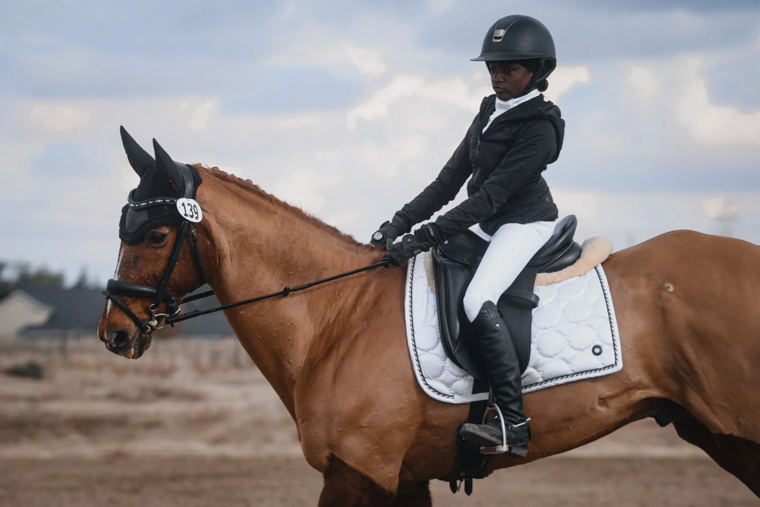 A woman wearing a black helmet, black riding jacket, and white riding pants sitting on a brown horse with a white saddle pad during daytime. The background shows a cloudy sky and an open outdoor riding area.