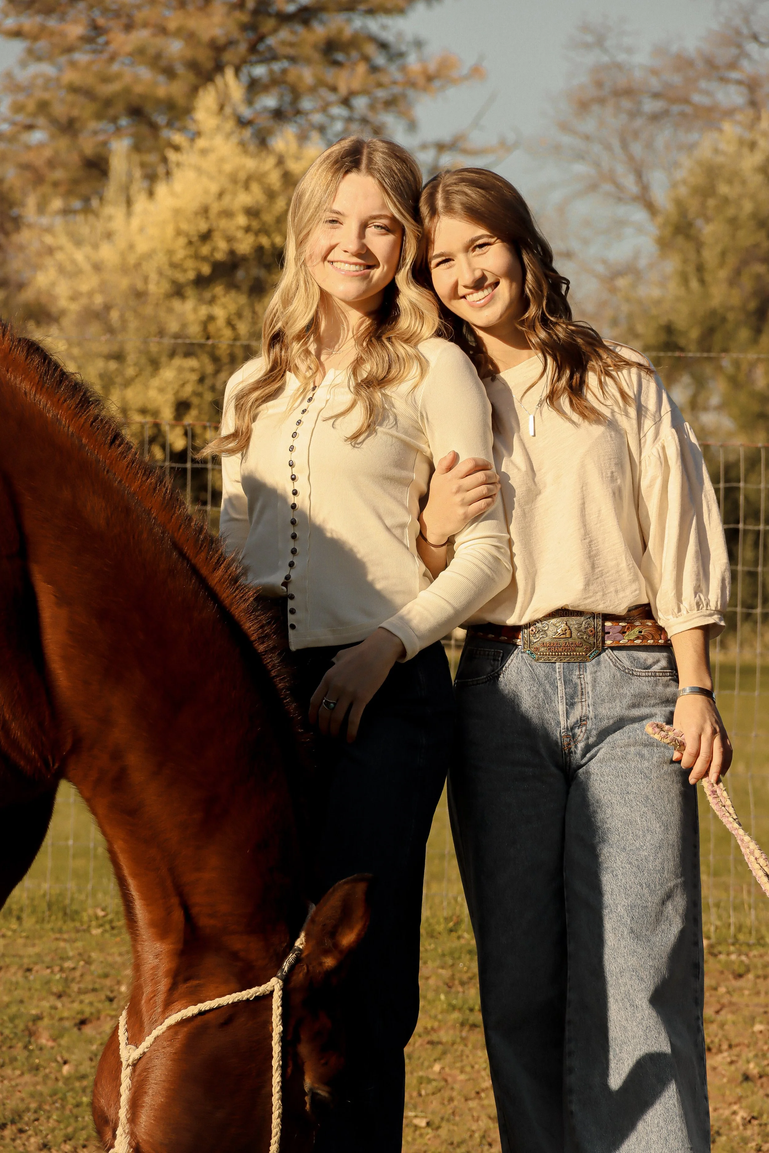Two young women stand outdoors smiling, one with blonde hair and the other with brown hair, holding a rope attached to a brown horse with a braided mane, in front of a metal fence and trees with autumn leaves.