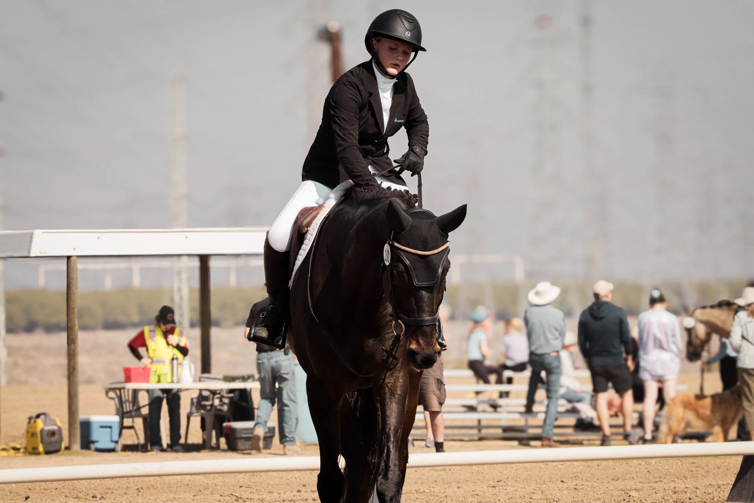 A young girl in riding gear, including a helmet, riding a black horse in an outdoor equestrian arena with people watching in the background.