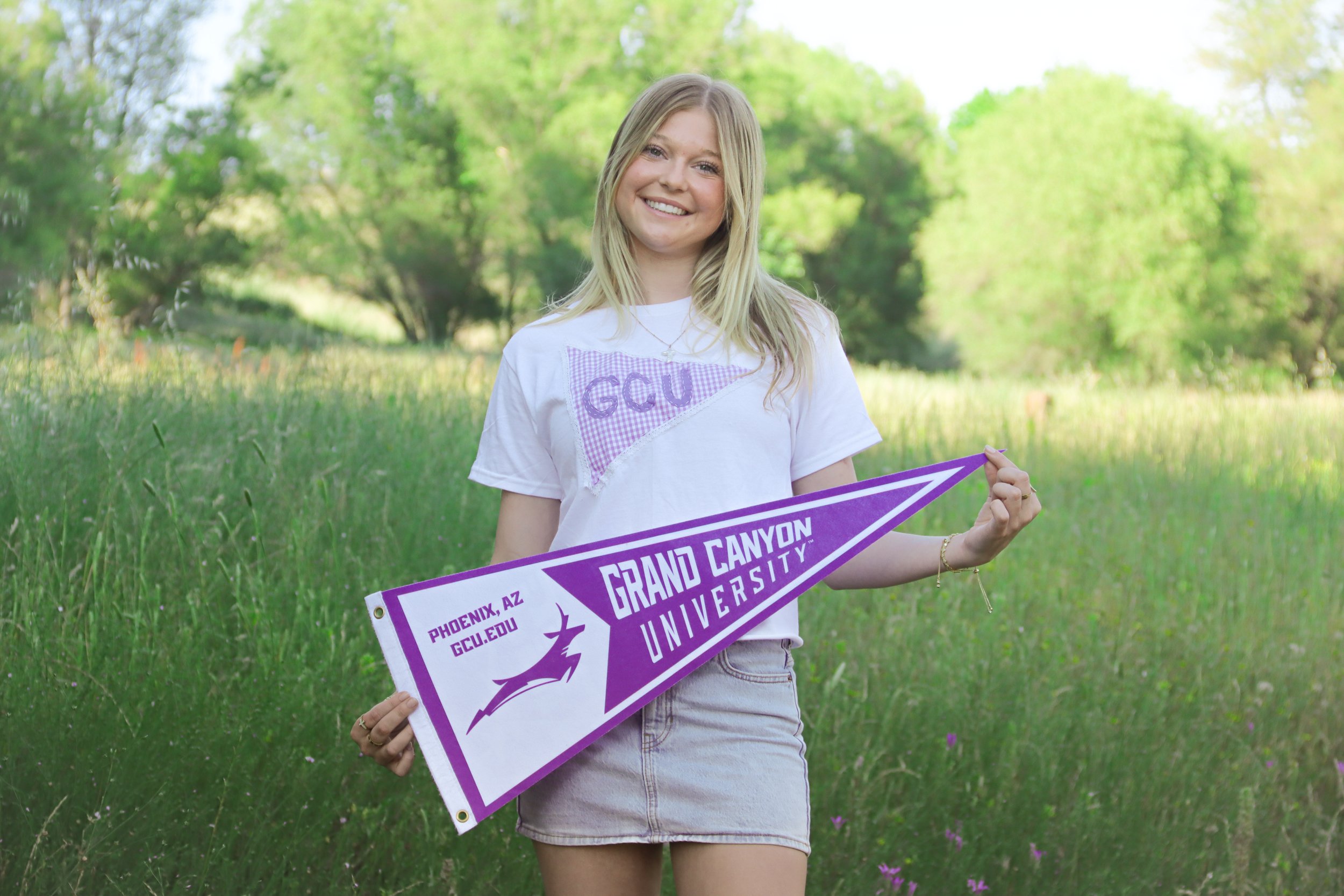 A young woman with blonde hair smiling outdoors in a lush green park, holding a purple and white University of Arizona Grand Canyon pennant. She is wearing a white T-shirt with purple lettering, and the background features trees and grass.