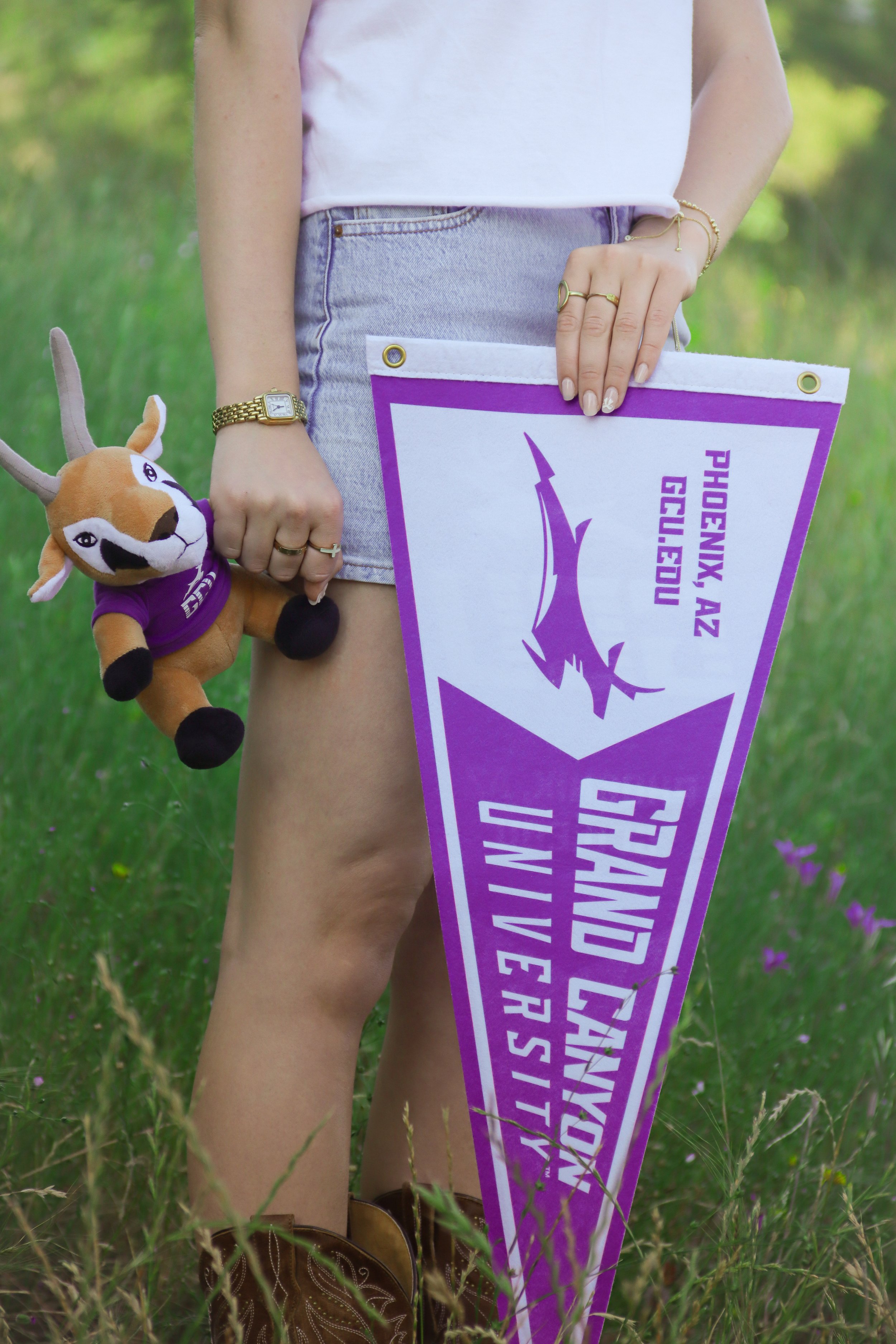 Part of a person standing outdoors on grass with purple and white flag and a stuffed antelope toy, wearing denim shorts and a light pink top, accessorized with jewelry and a watch.