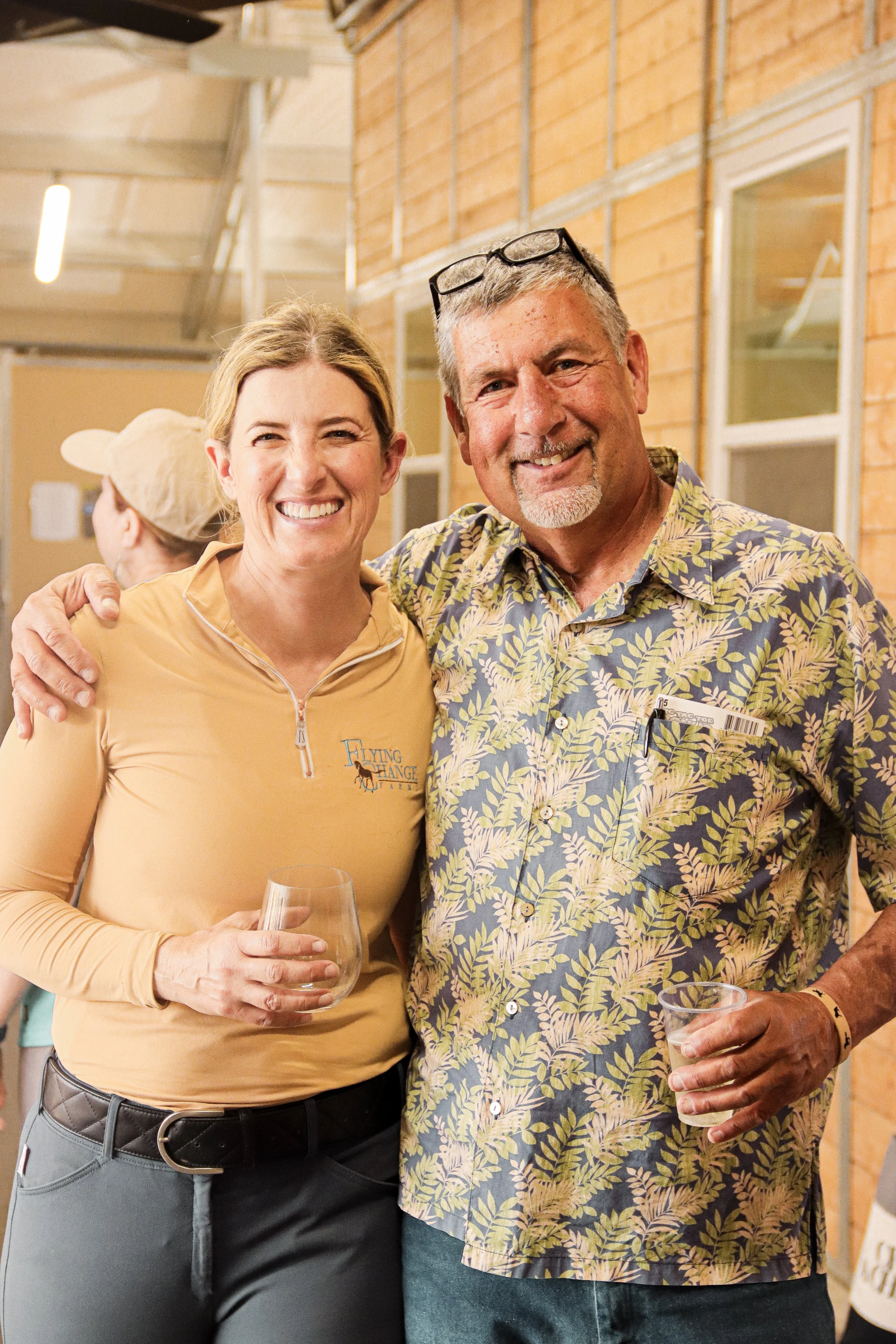 A woman and a man smiling and posing together at an indoor event. The woman is wearing a yellow long-sleeve shirt with a logo and is holding a wine glass. The man is wearing a floral shirt with glasses on his head and is holding a cup.