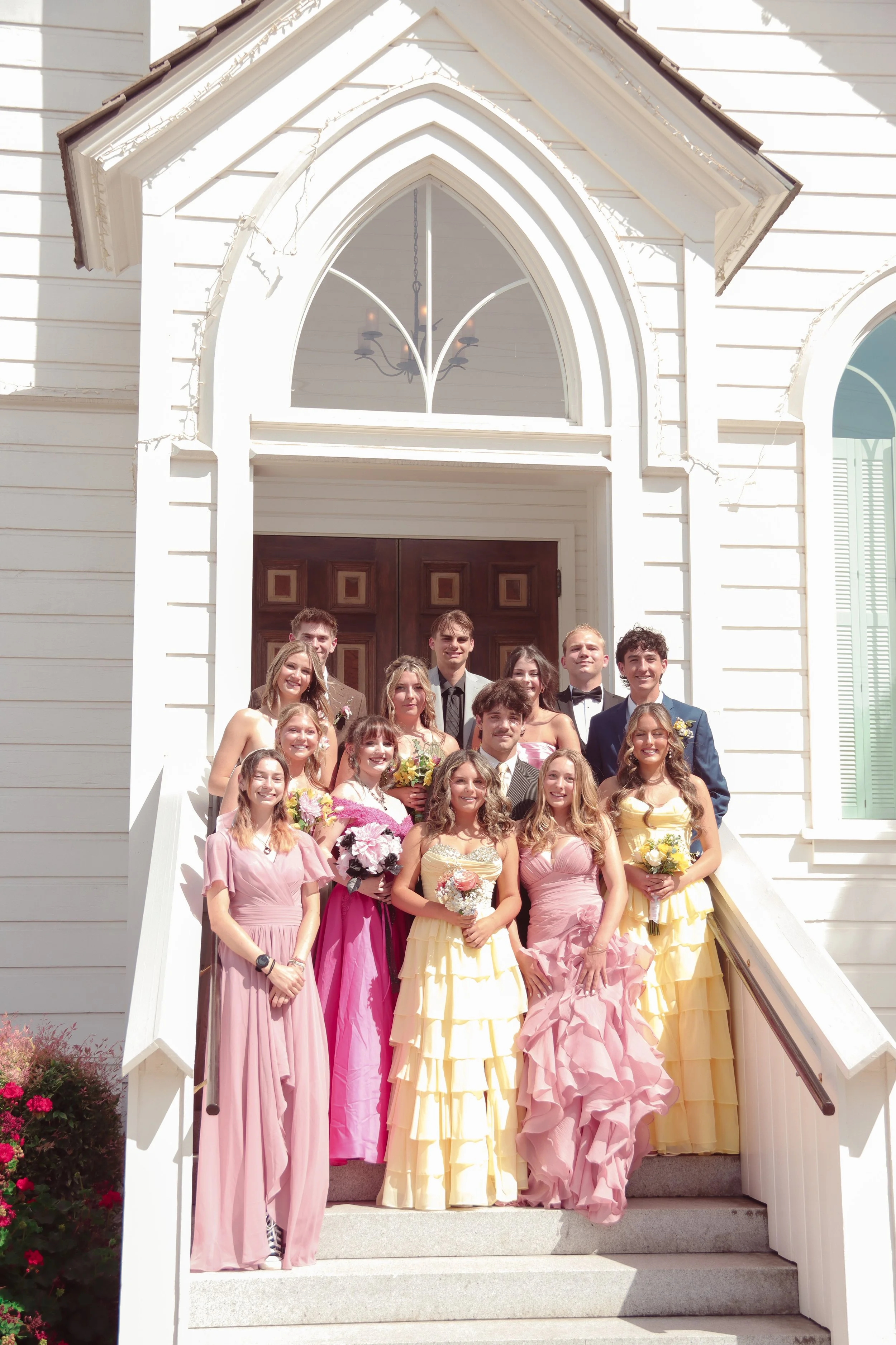 Group of young people in formal attire standing on the steps of a white church with a Gothic arch window above the entrance during daytime.