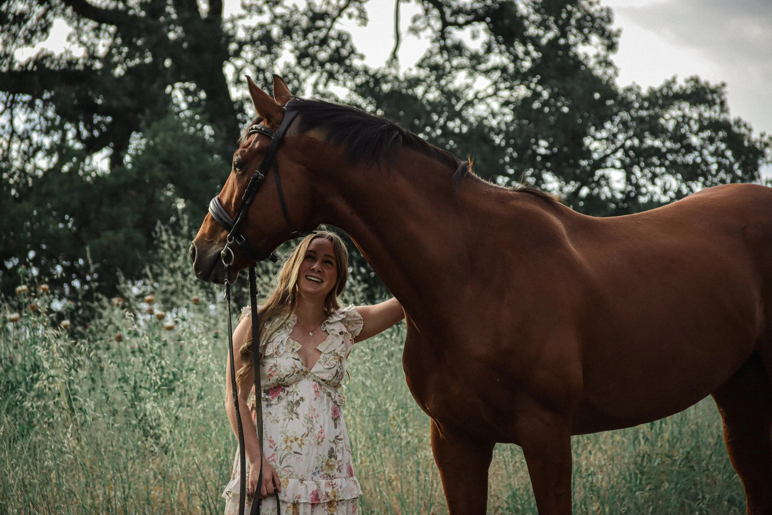 A woman with long blonde hair, wearing a floral dress, smiling and holding a brown horse's bridle outdoors in a green field with trees in the background.