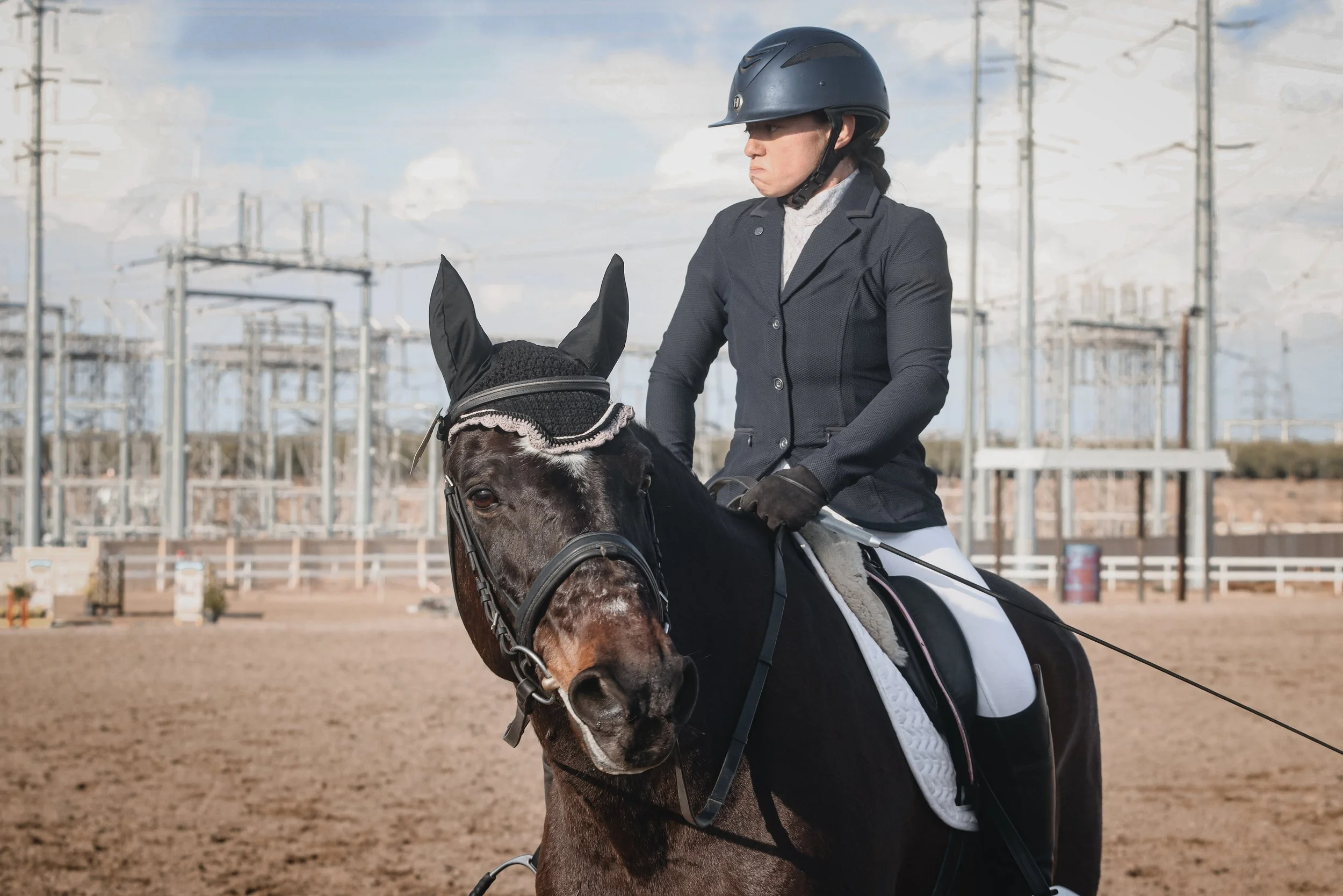 A person wearing a black riding helmet and riding a brown horse with a black mane in an outdoor horse riding arena with electrical power lines and towers in the background.