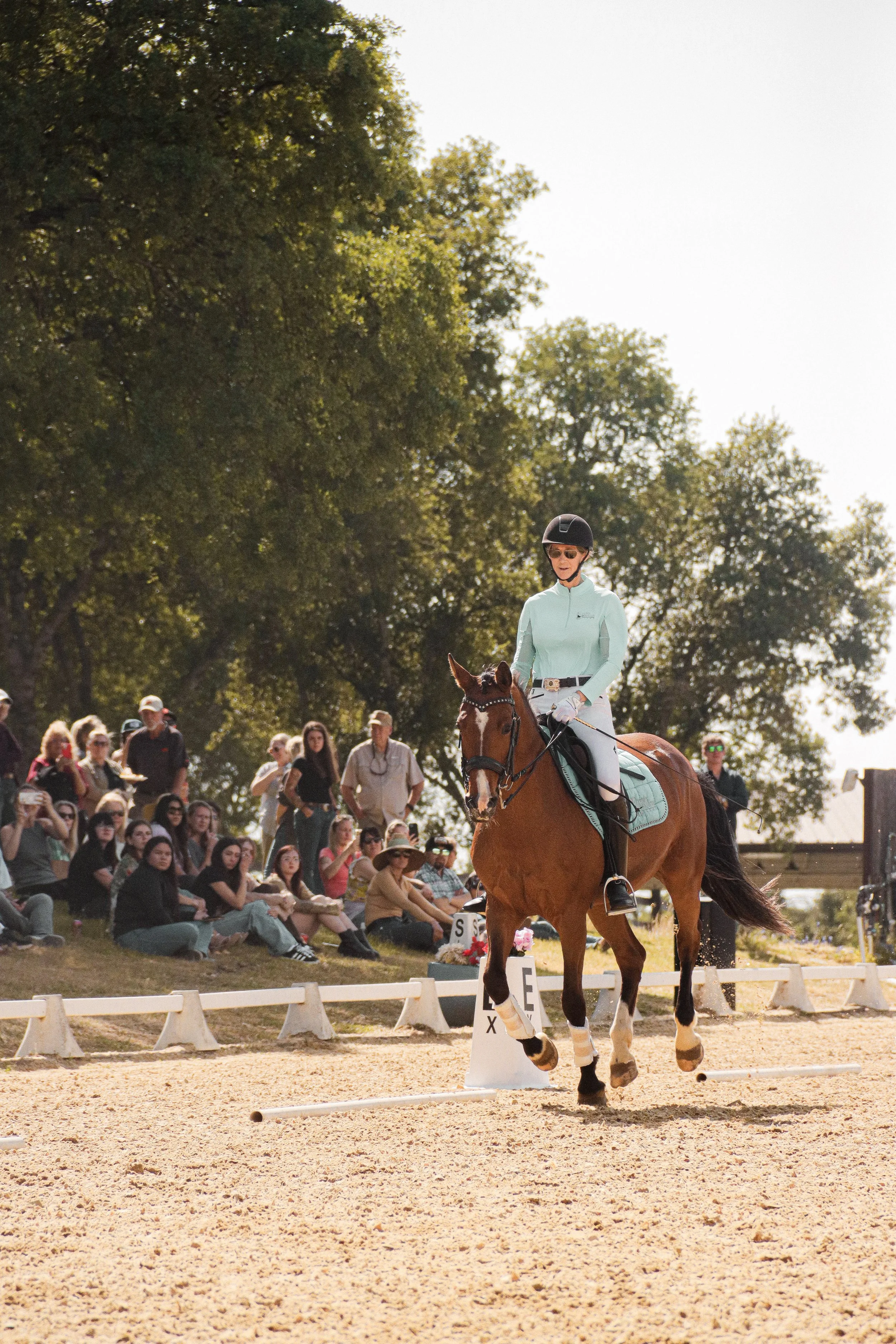 A woman riding a horse during an equestrian event, with spectators watching in the background on a sunny day.