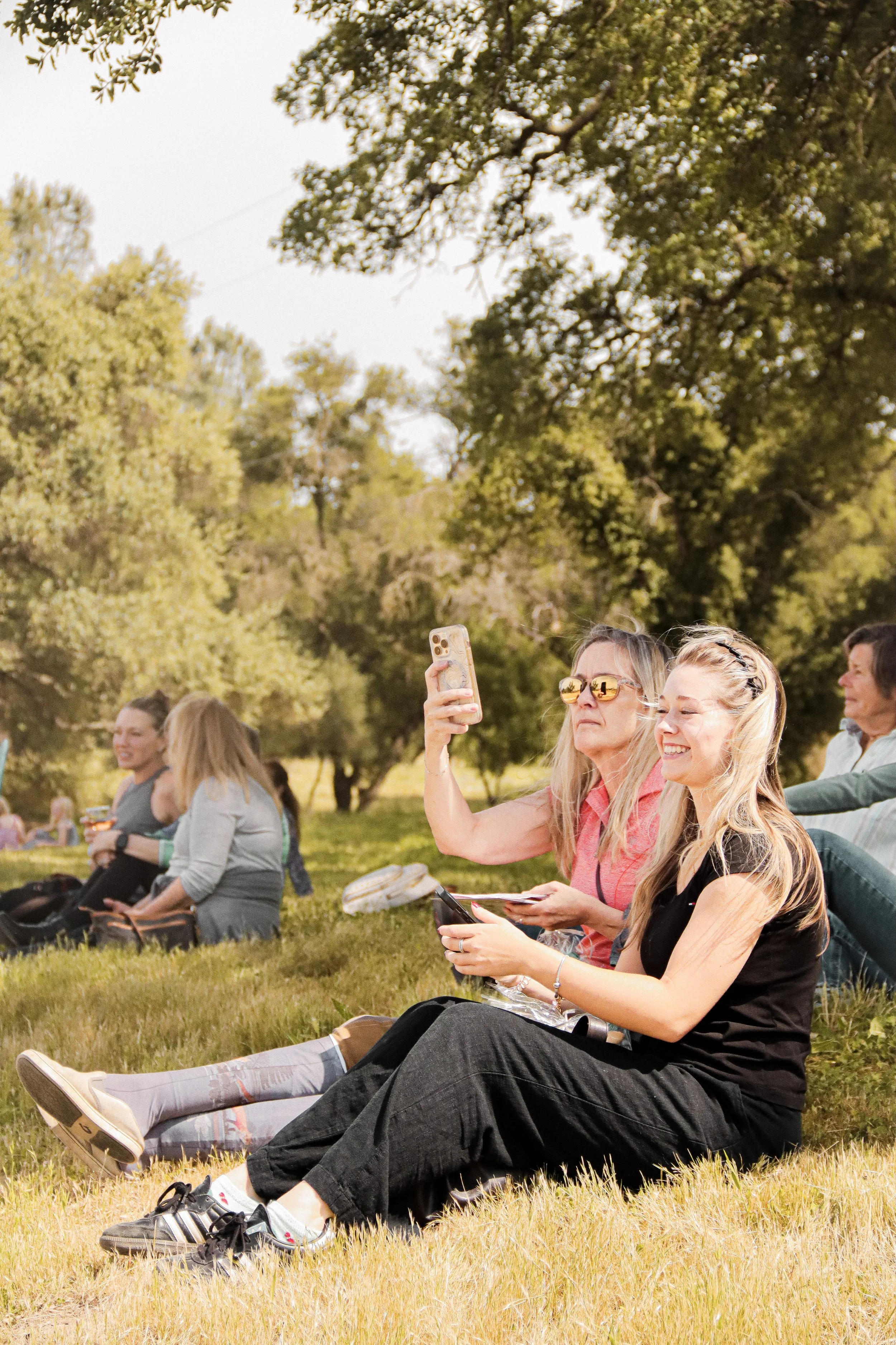 Group of people sitting on grass outdoors, some taking selfies and others relaxing at a park with trees in the background.