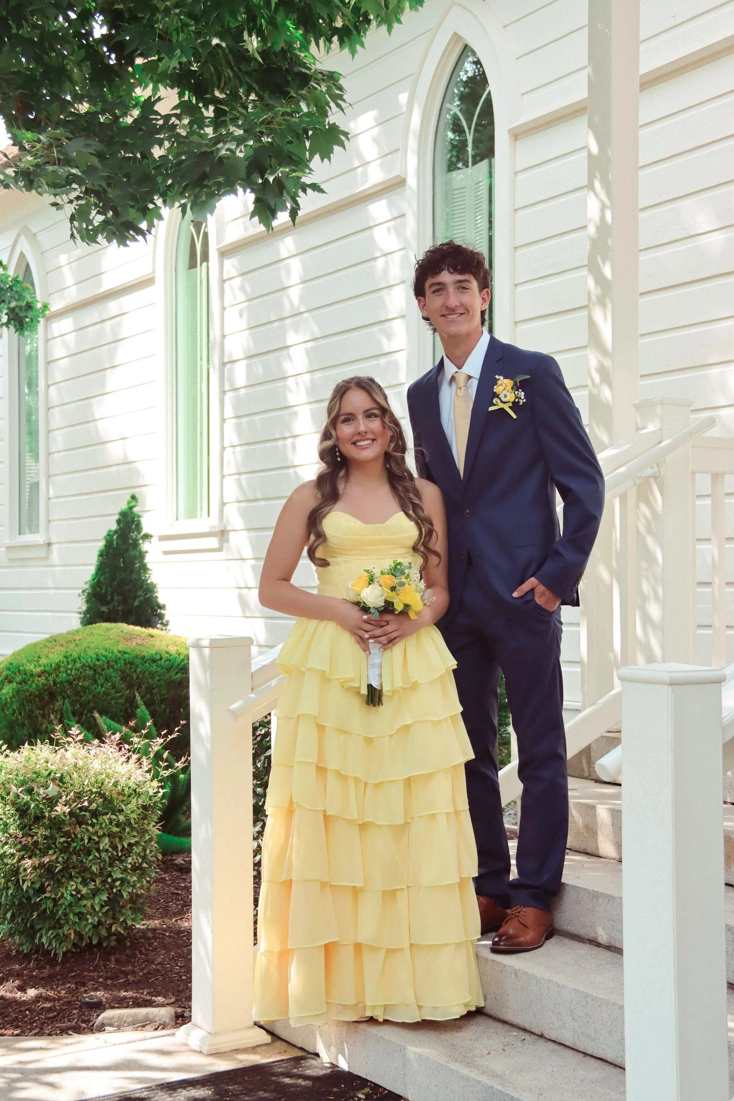 A young woman in a yellow tiered dress holding a bouquet, standing on the steps of a white house with a young man in a navy suit and yellow tie beside her. They are smiling outdoors on a sunny day, with greenery in the background.