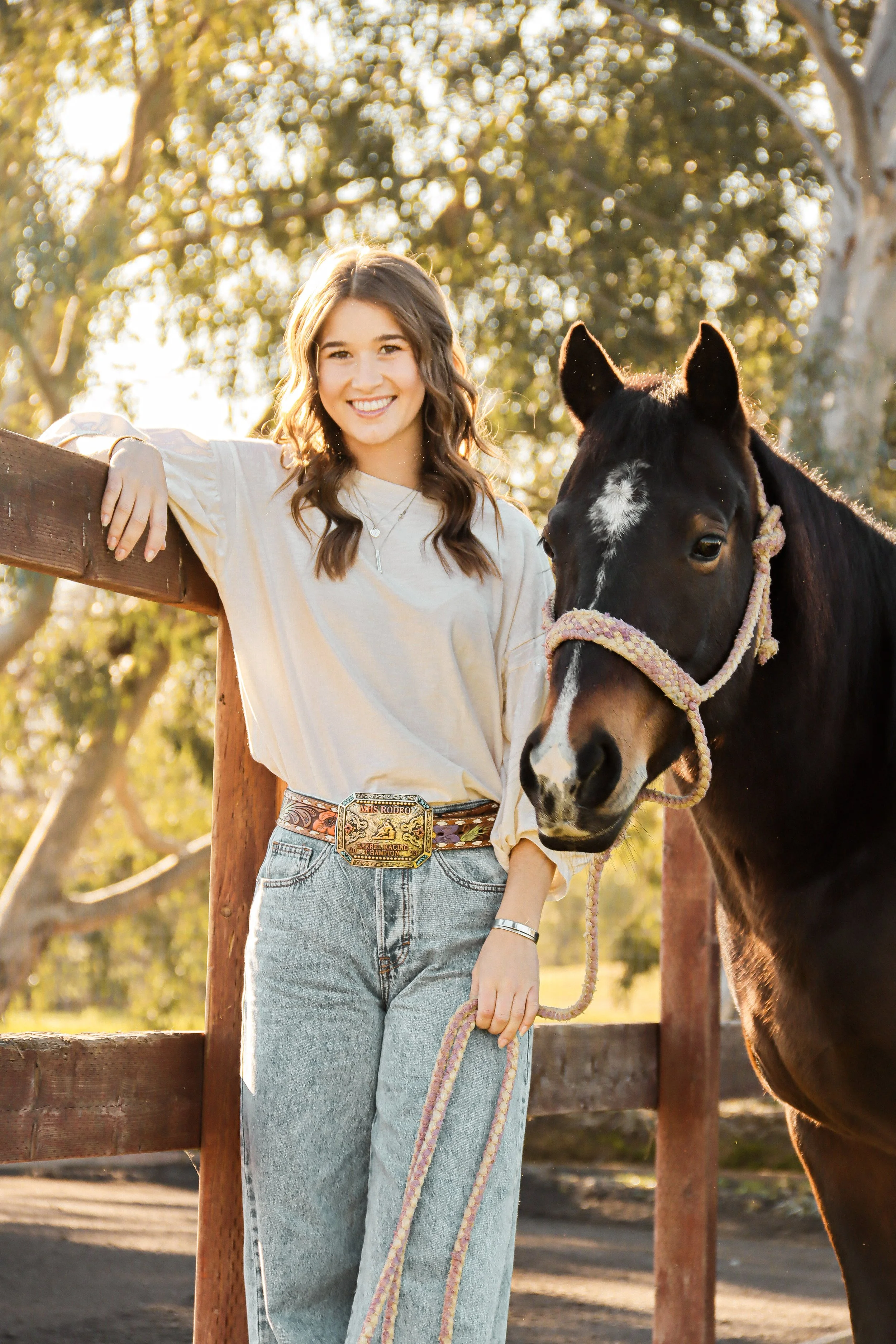 A young woman with long brown hair, smiling, standing beside a dark horse with a white star marking on its forehead, outdoors during golden hour with trees and sunlight in the background.