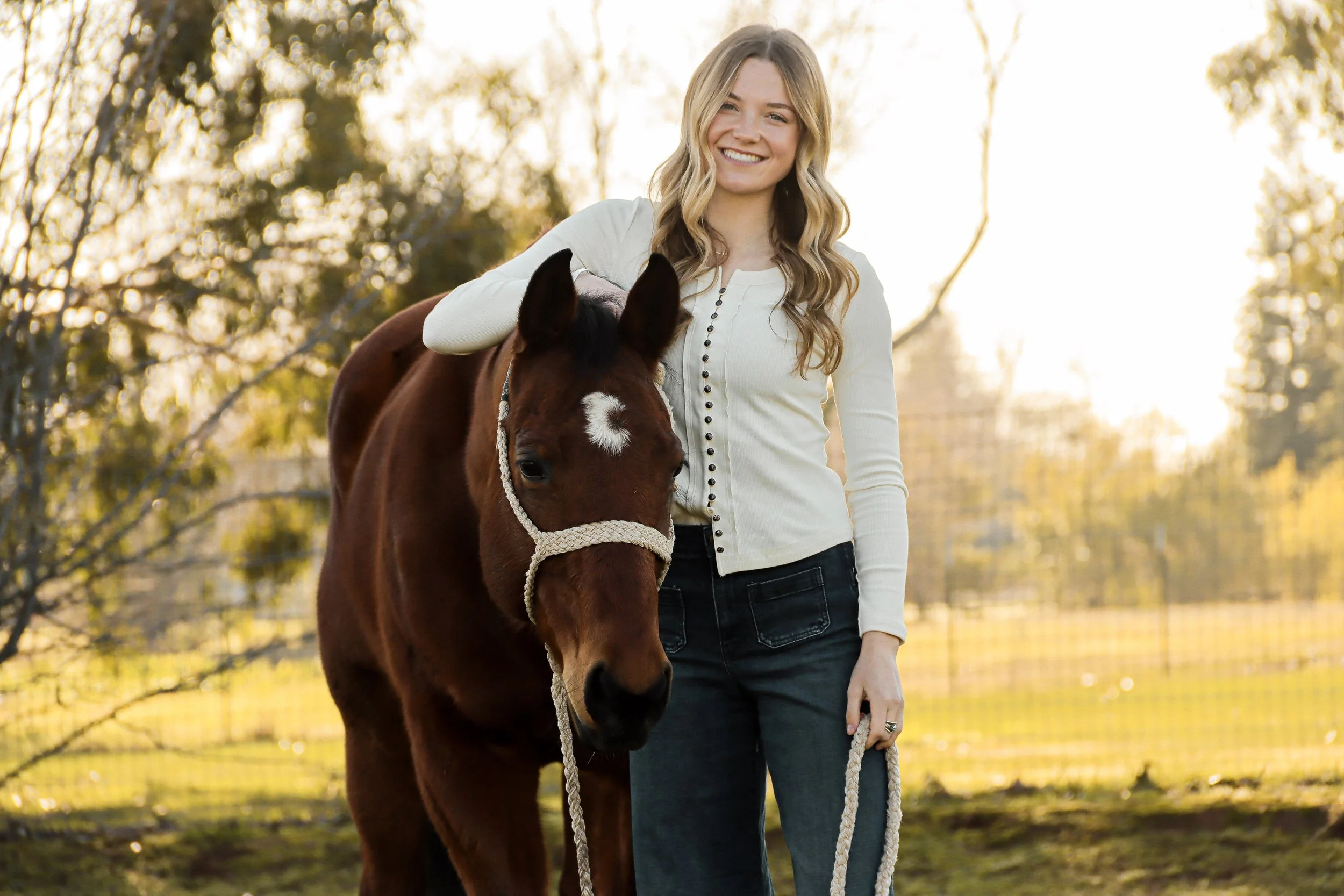 A young woman with long blonde hair smiling while standing outdoors holding a brown horse with a white star on its forehead during sunset.