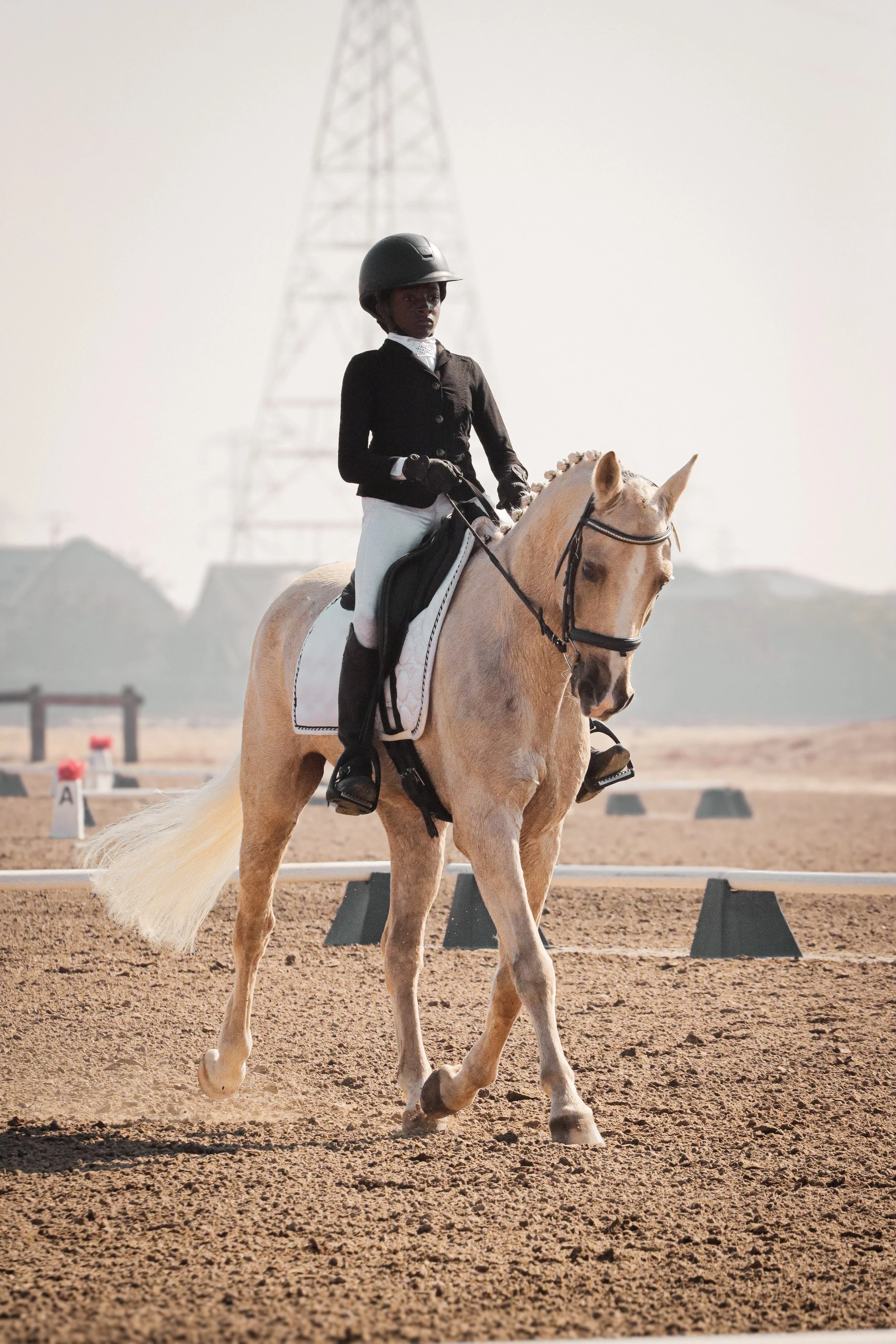 A person riding a light-colored horse during an equestrian event on a dirt arena with markers and a distant power line tower.