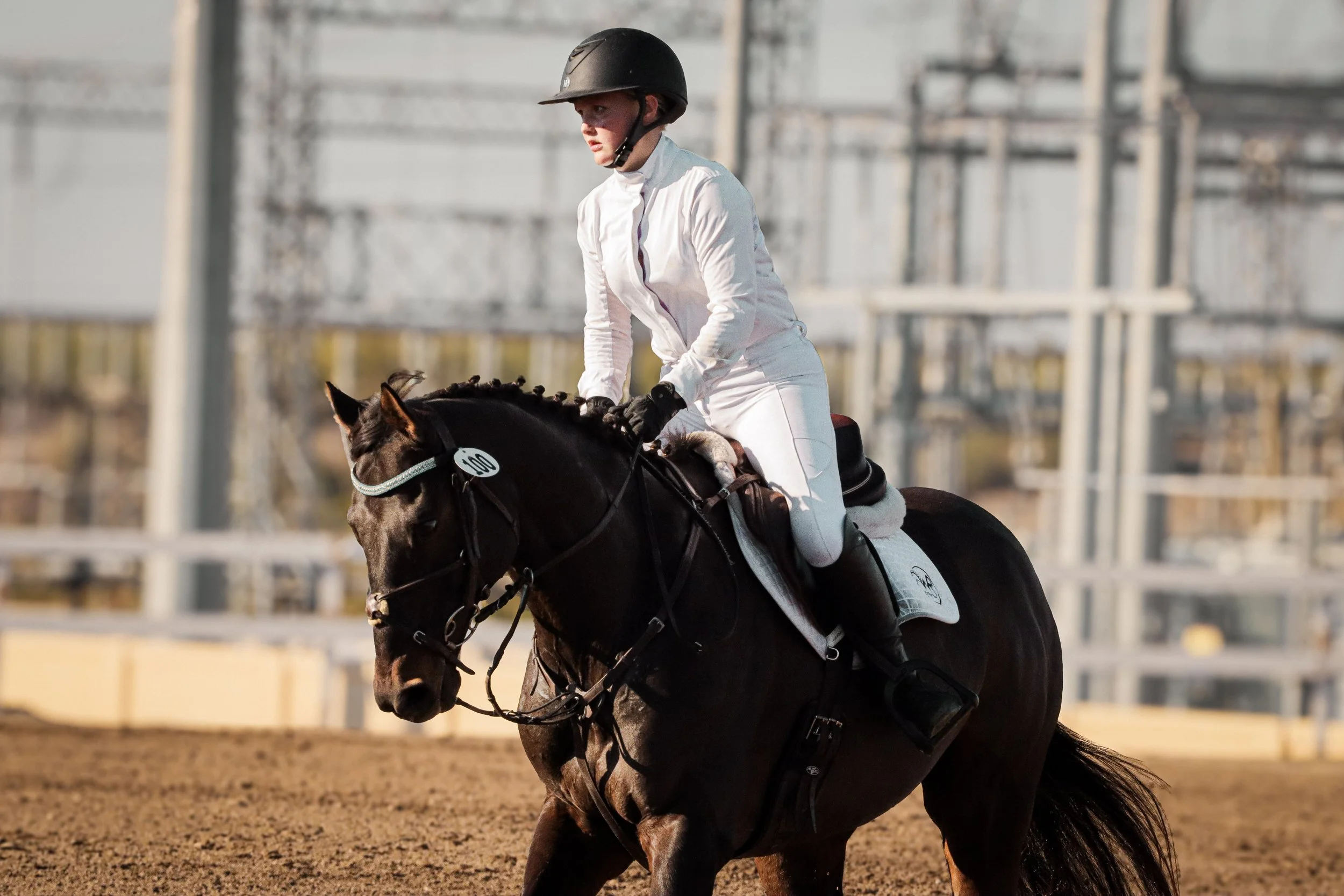 A female equestrian rider in white riding attire and a black helmet riding a dark brown horse on an outdoor dirt track.