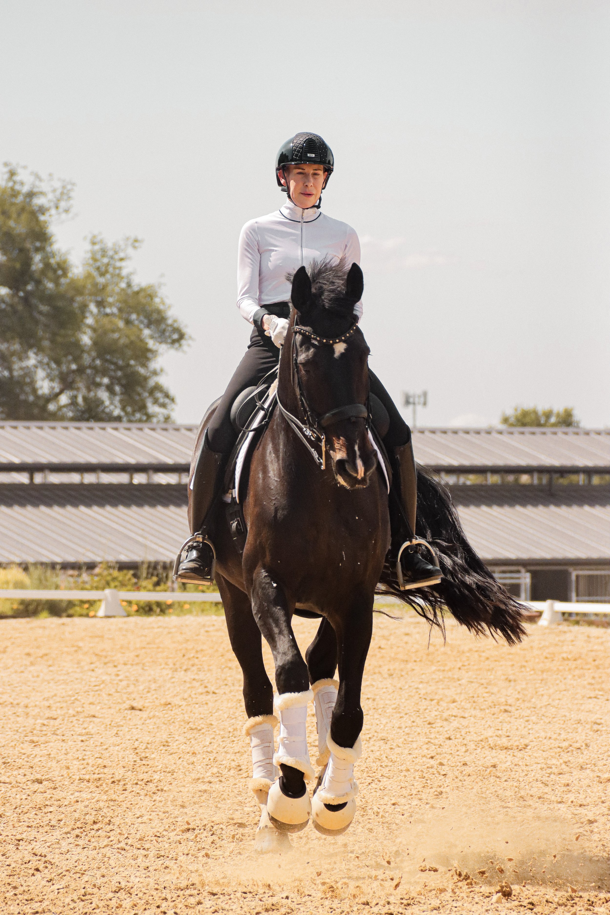Person riding a horse in an outdoor riding arena, wearing riding gear including a helmet, gloves, and equestrian boots, with dust being kicked up from the ground.