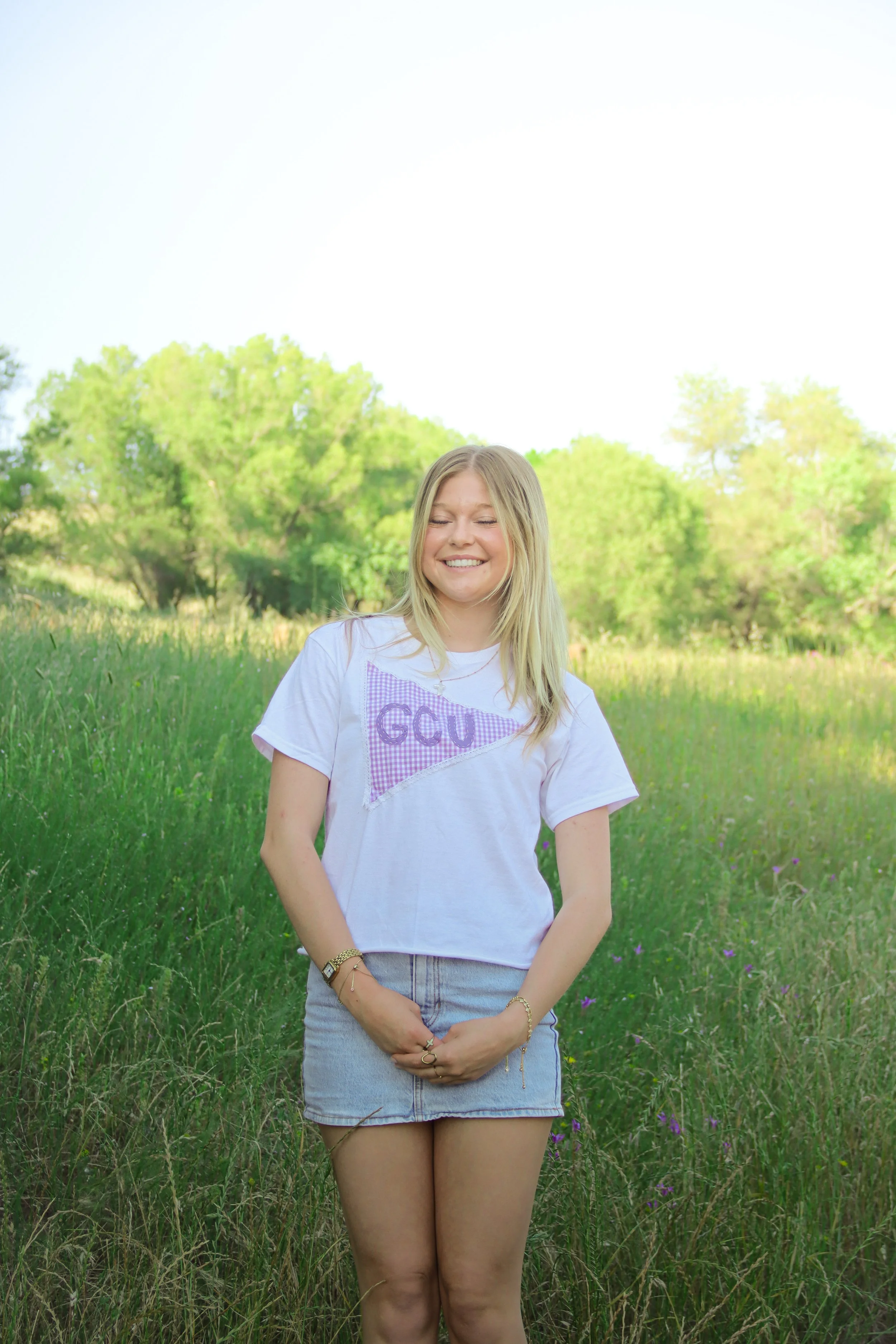 A young woman standing outdoors in a grassy field with green trees in the background, smiling with her eyes closed, wearing a white t-shirt and a denim skirt.
