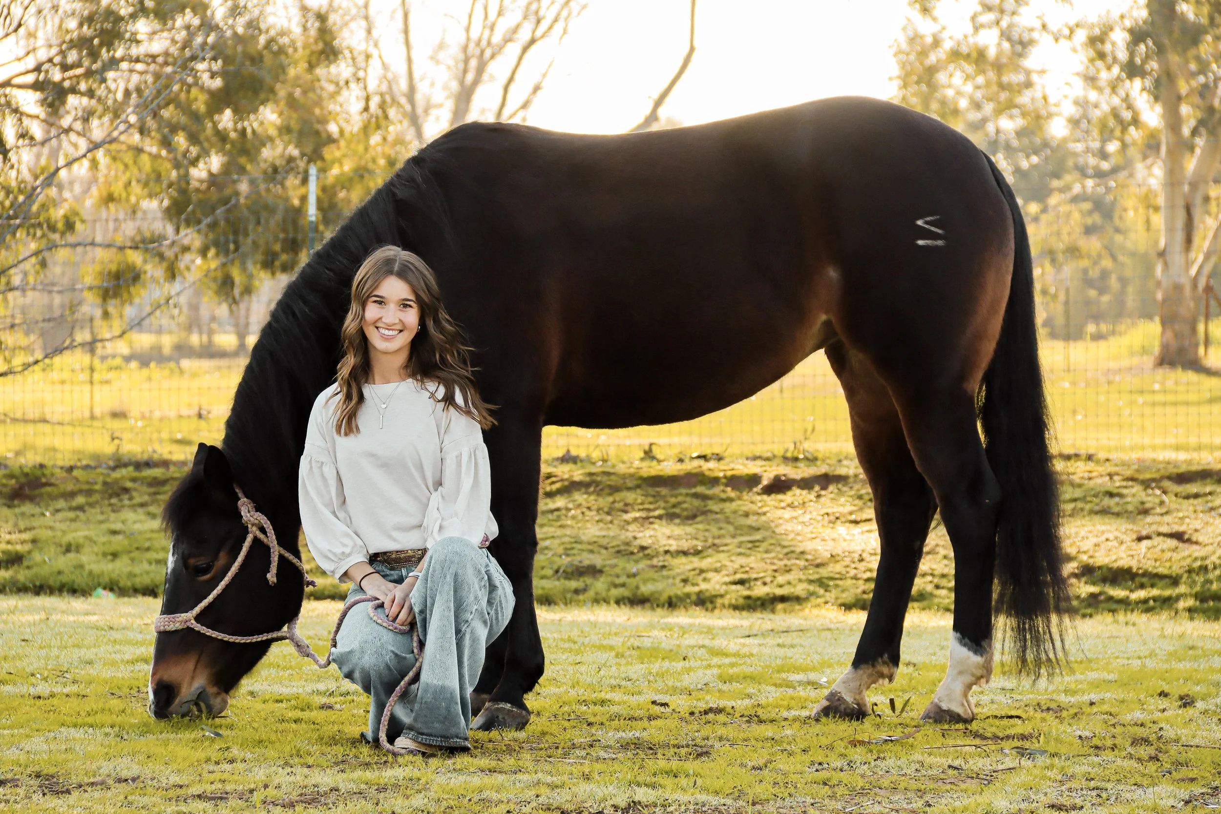 A smiling young woman with long brown hair sitting on the grass next to a large black and brown horse with a rope halter, in a sunlit outdoor setting with trees and a fence in the background.