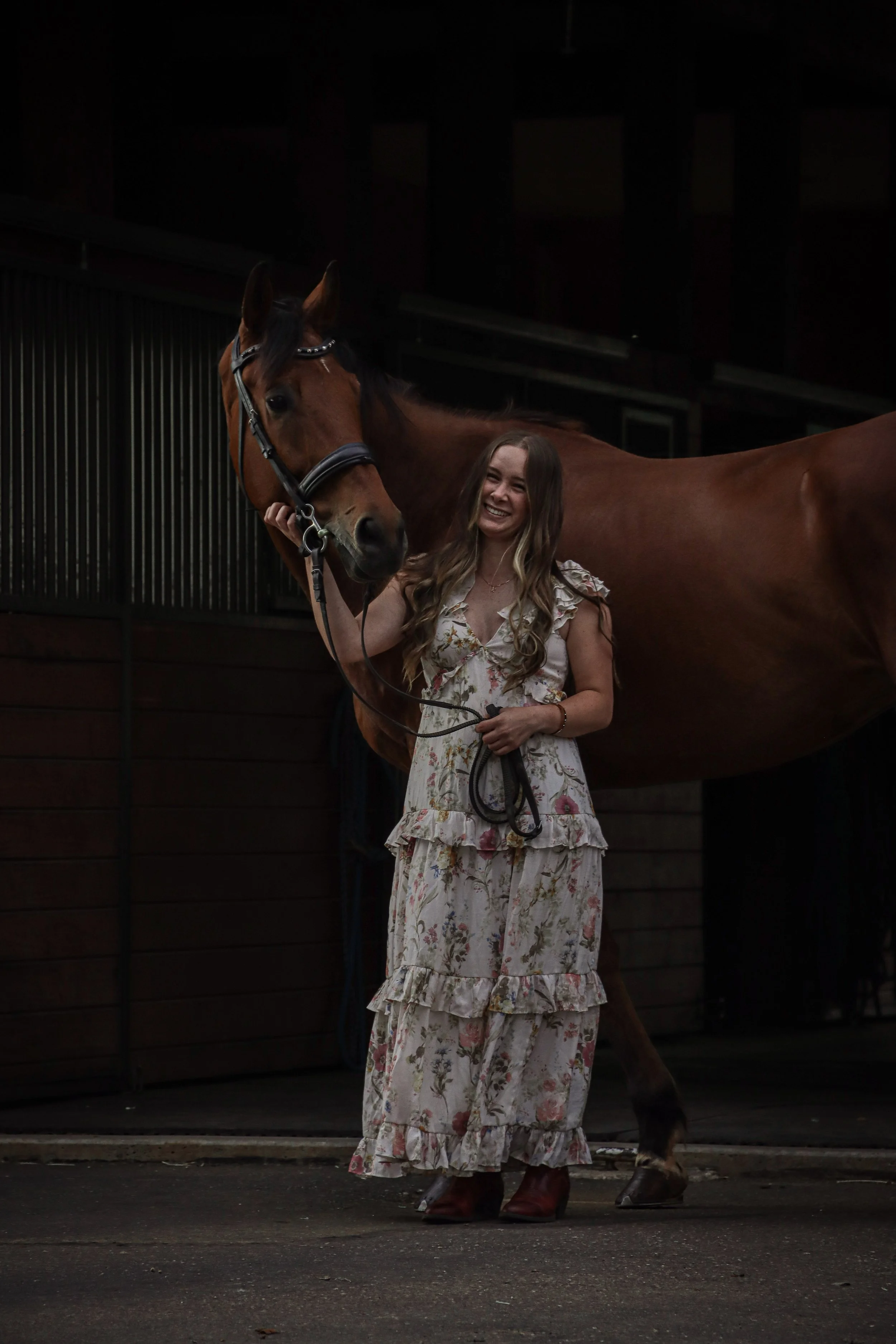 A woman in a floral dress and red cowboy boots standing beside a brown horse in a dark stable or barn.