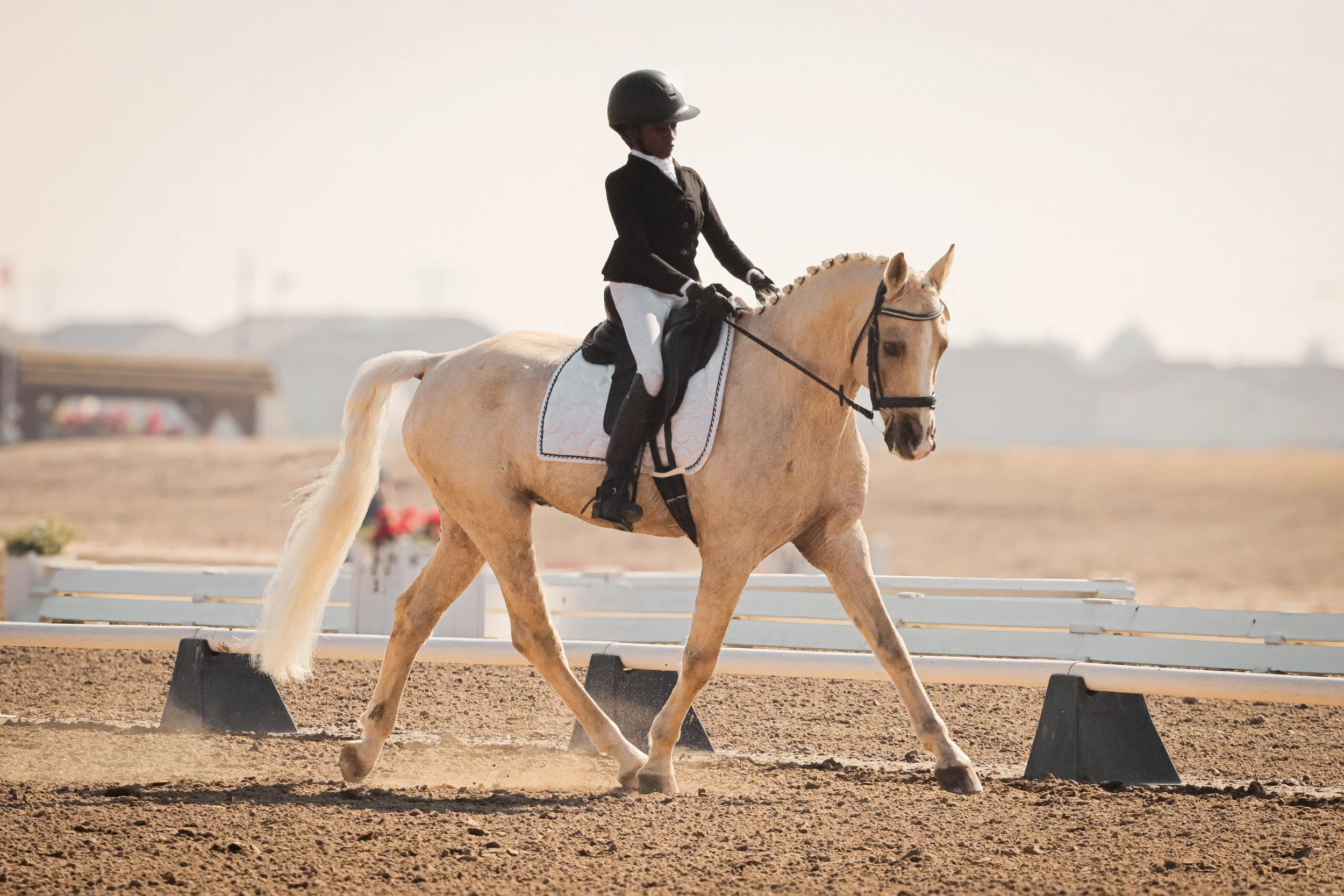 A person riding a light-colored horse on a dirt riding arena during daytime.